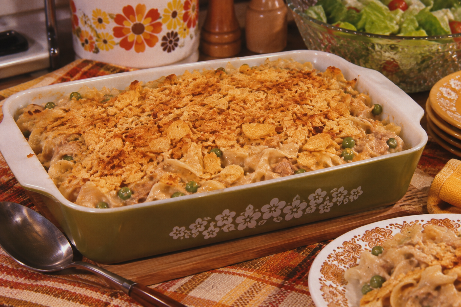 A baked casserole topped with crunchy crumbs sits in a green floral dish on a plaid tablecloth, next to a serving spoon, a salad, plates, and salt and pepper shakers.