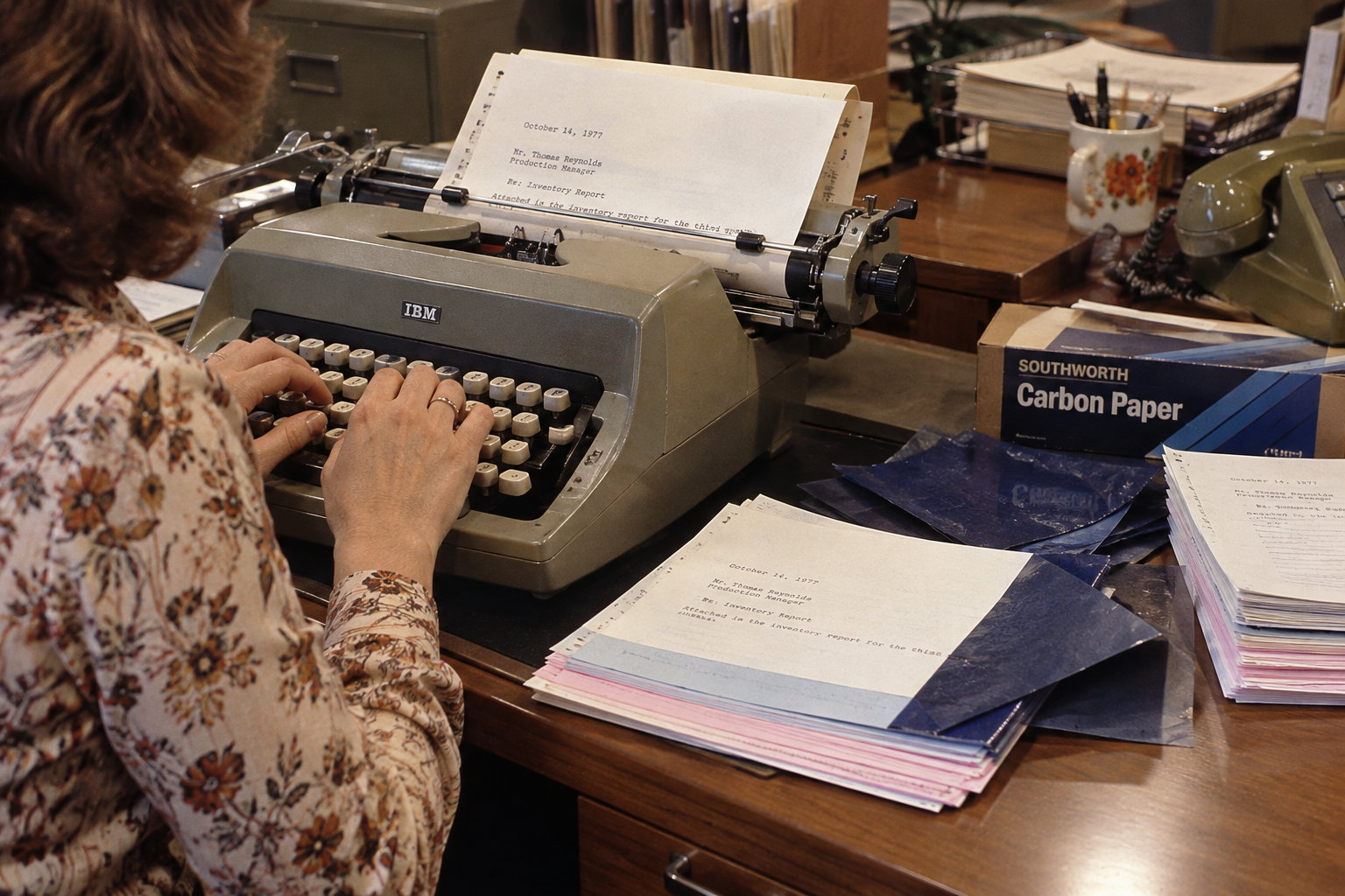 A person typing on an IBM electric typewriter, with a stack of carbon paper and typed reports on the desk. Office life before email.