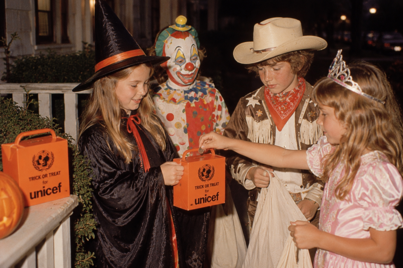 Four children in Halloween costumes standing on a porch, with one placing a coin into an orange UNICEF collection box.