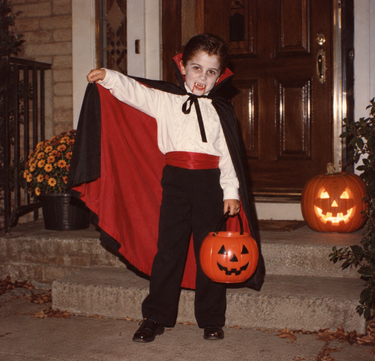 A young child in a vampire costume with a red-lined cape, plastic fangs, and a pumpkin-shaped candy bucket stands on a porch in front of a lit jack-o’-lantern.