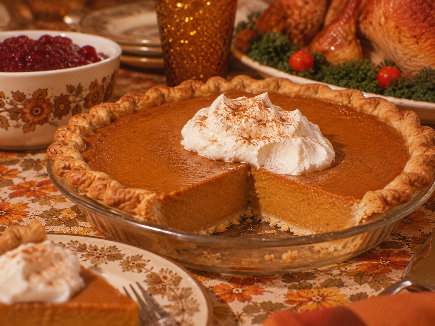 A pumpkin pie topped with whipped cream and cinnamon, with a slice removed, served on a table with a roasted turkey and bowl of cranberry sauce.