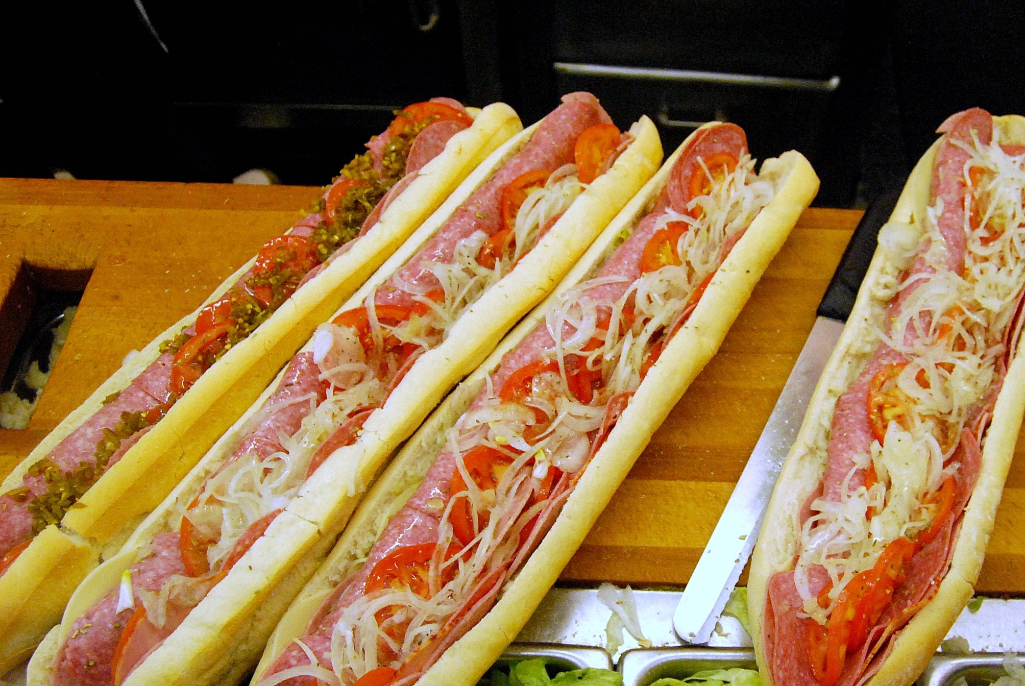 Restaurants Across The USA Known For Next-Level Hoagies, Four long sub sandwiches on a wooden counter, topped with salami, sliced tomatoes, onions, and relish.