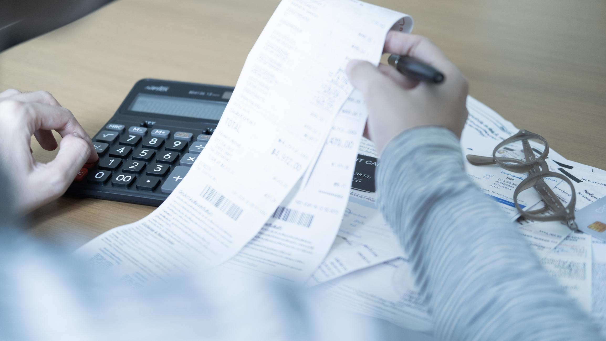 A person using a calculator at a desk while reviewing a long paper receipt of cruise ship charges.
