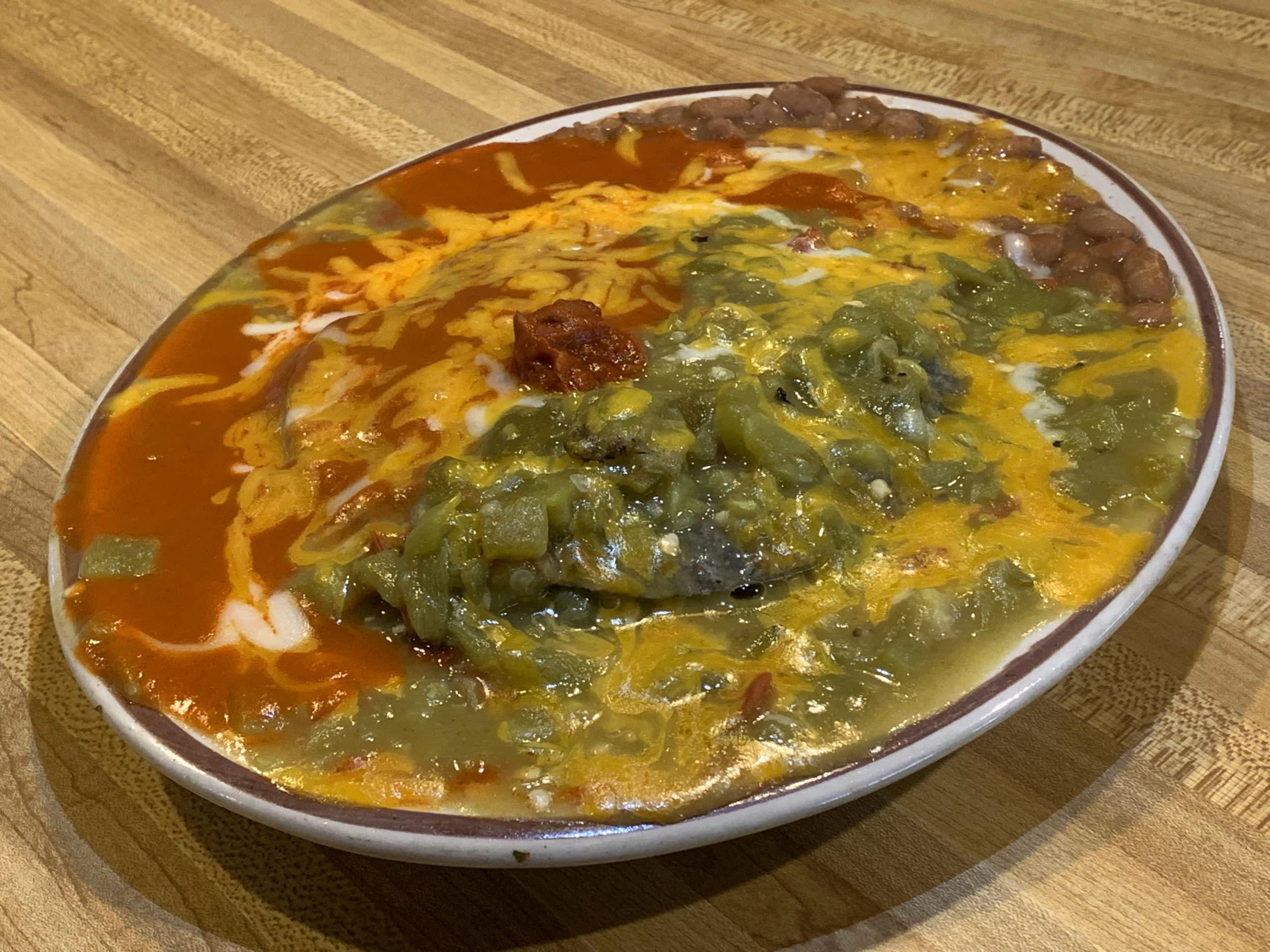 A plate of food reflecting American dining traditions, featuring red and green chile sauces, melted cheese, and a side of pinto beans.