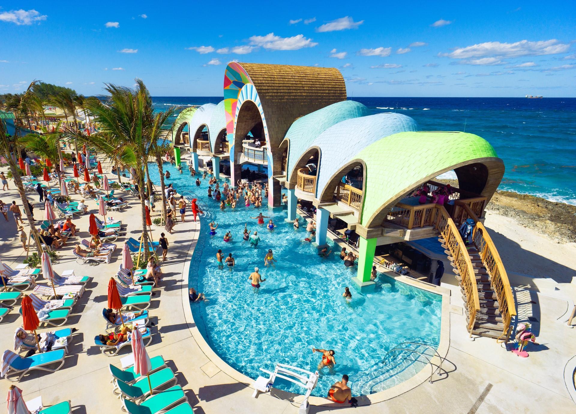 Aerial view of a coastal swimming pool complex with colorful, rounded architecture and people swimming, surrounded by lounge chairs and palm trees along a beach.