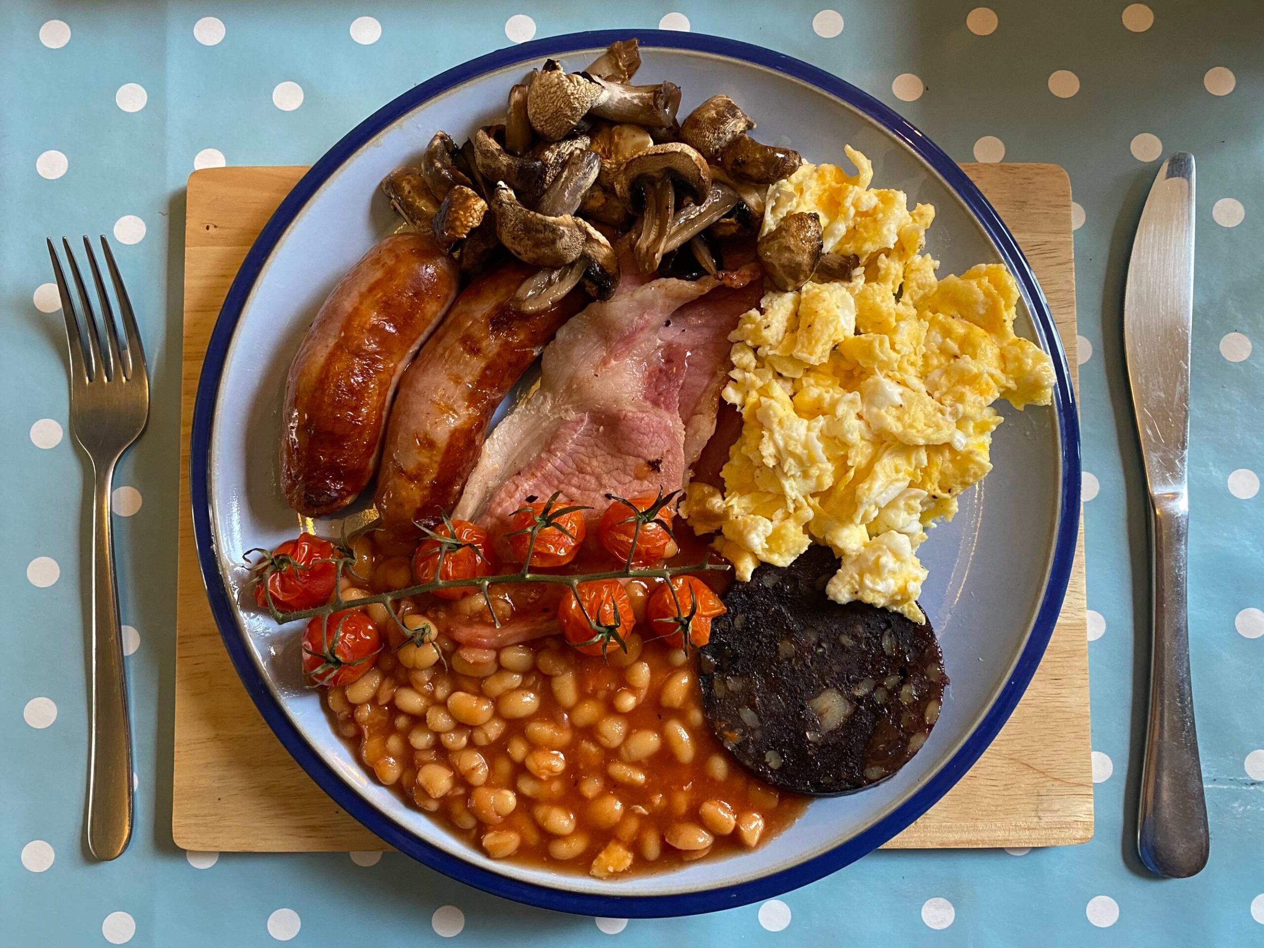 A breakfast plate with sausages, bacon, scrambled eggs, black pudding, baked beans, mushrooms, and cherry tomatoes on the vine, served with a fork and knife.