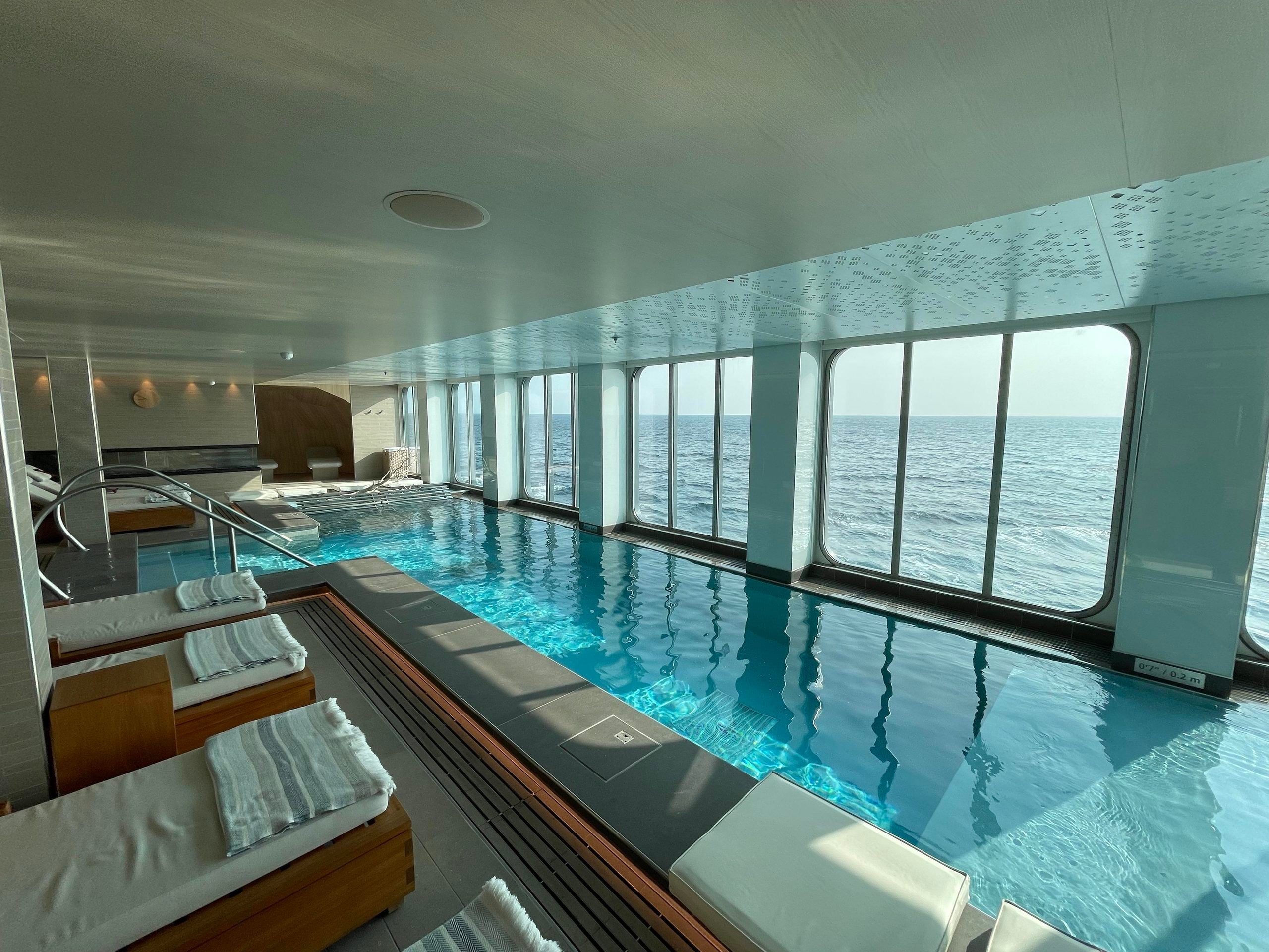 Indoor swimming pool with lounge chairs alongside floor-to-ceiling windows overlooking the ocean.