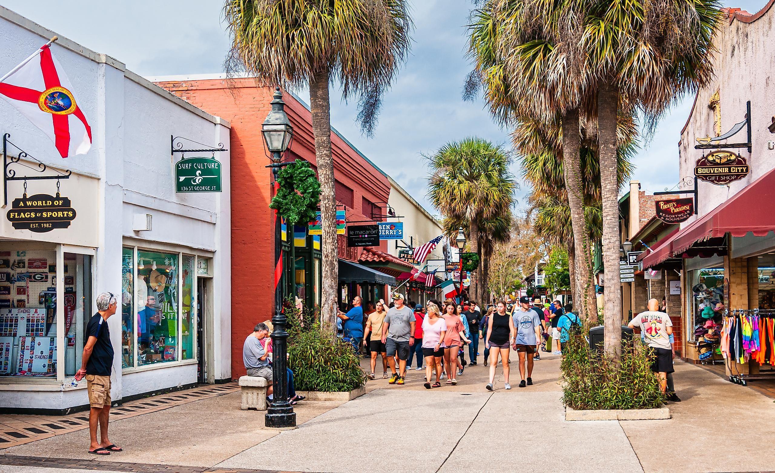 Pedestrians walk along a sunny, tree-lined street filled with shops, including a flag store, a souvenir shop, and a Ben & Jerry’s.