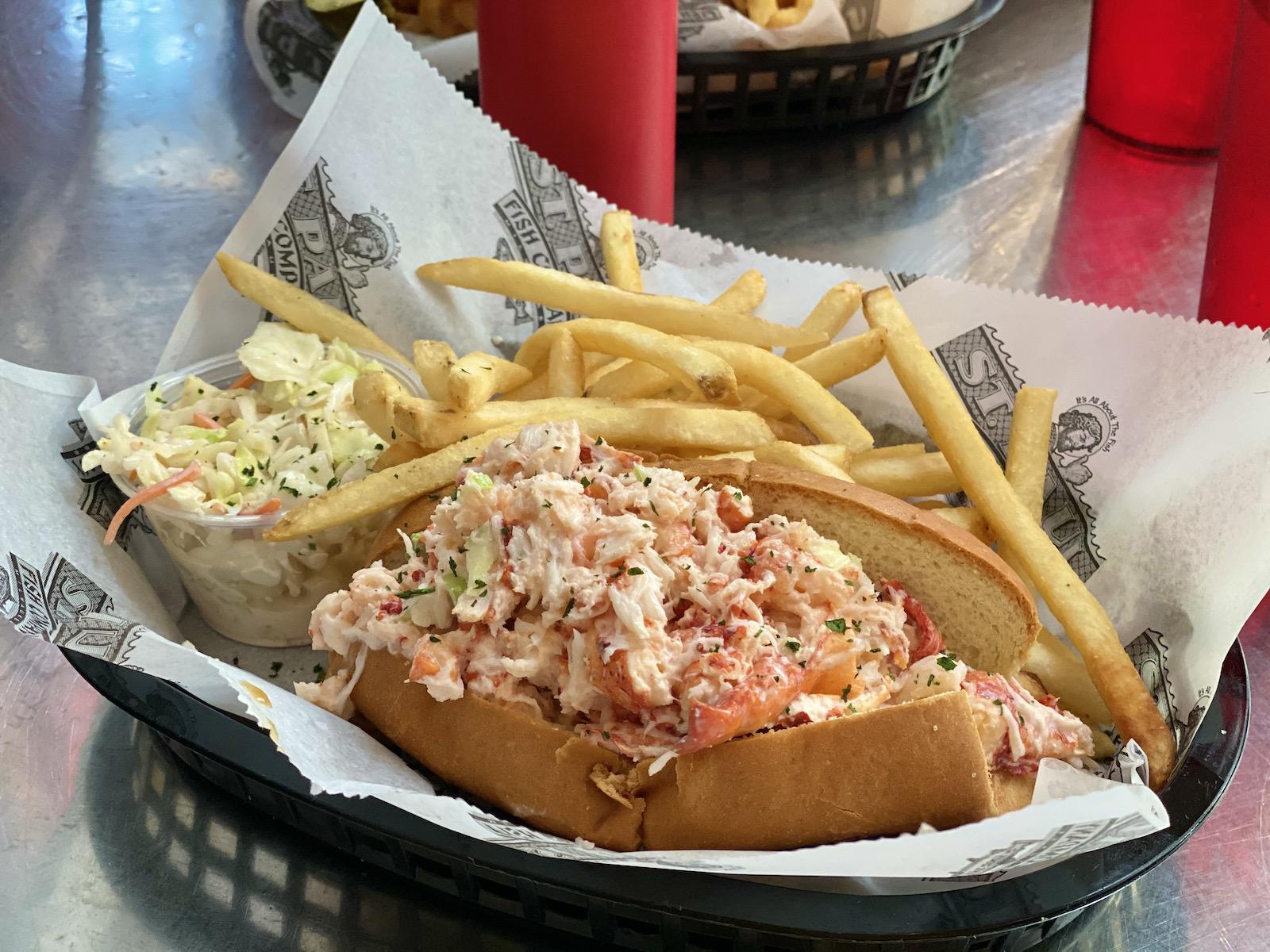 A lobster roll in a toasted bun served in a plastic basket with a side of coleslaw and french fries.