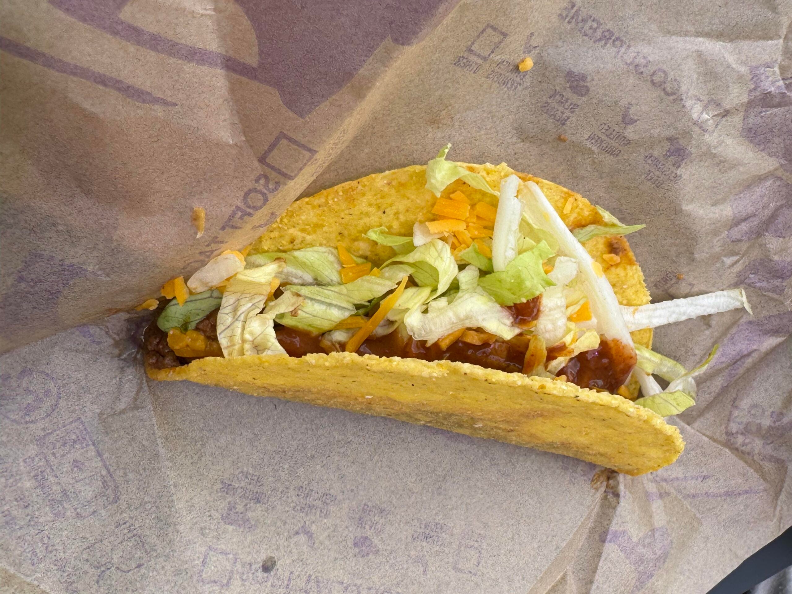 A crunchy yellow corn taco with seasoned meat, shredded cheese, and lettuce in a paper wrapper.