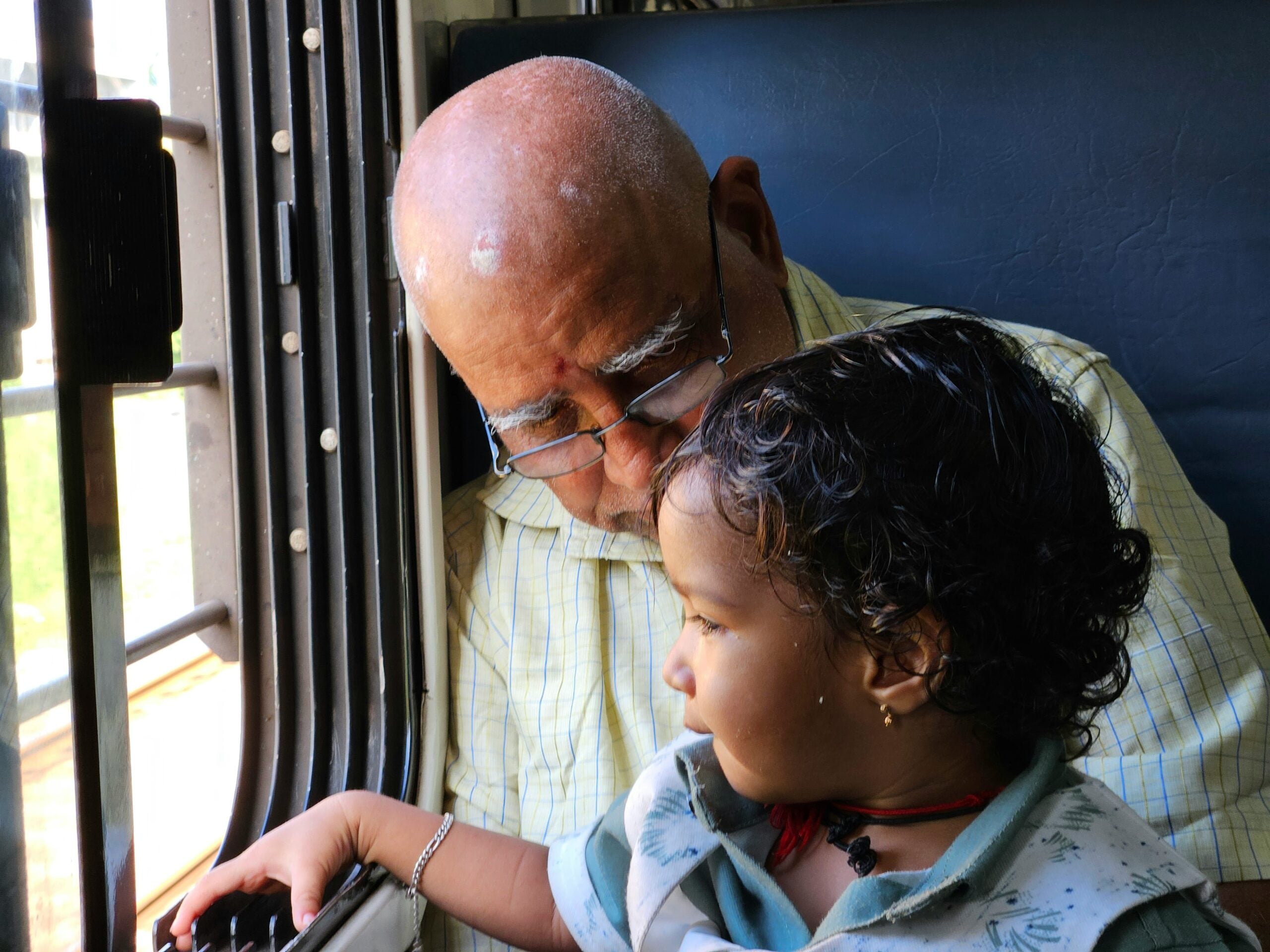 An elderly man with glasses and a young child sit together on a train, looking out the window. The man rests his head gently on the child, who is reaching toward the window bars.