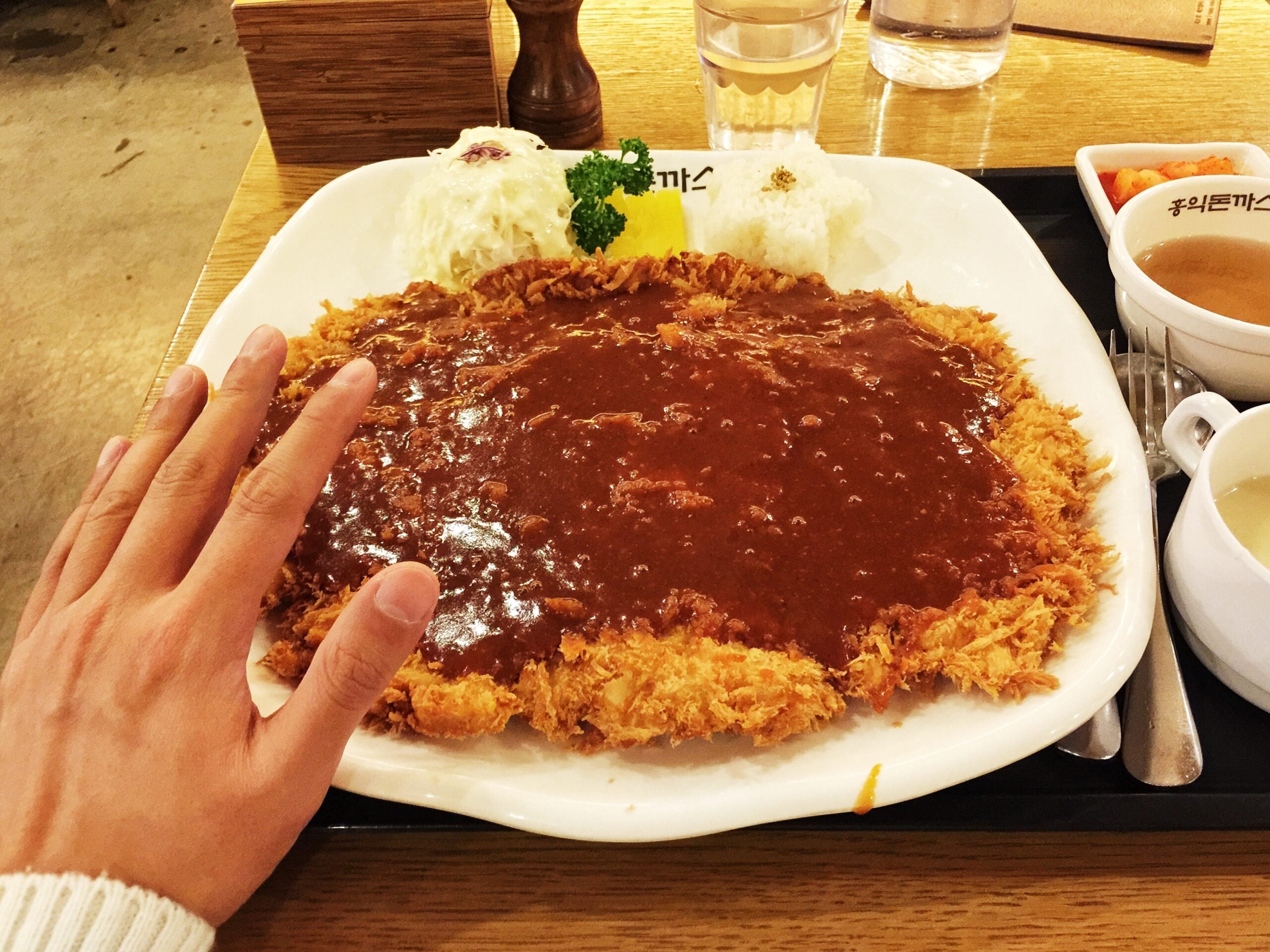 A large plate of breaded pork cutlet covered in brown sauce, served with a scoop of rice, shredded cabbage, and a side of soup.