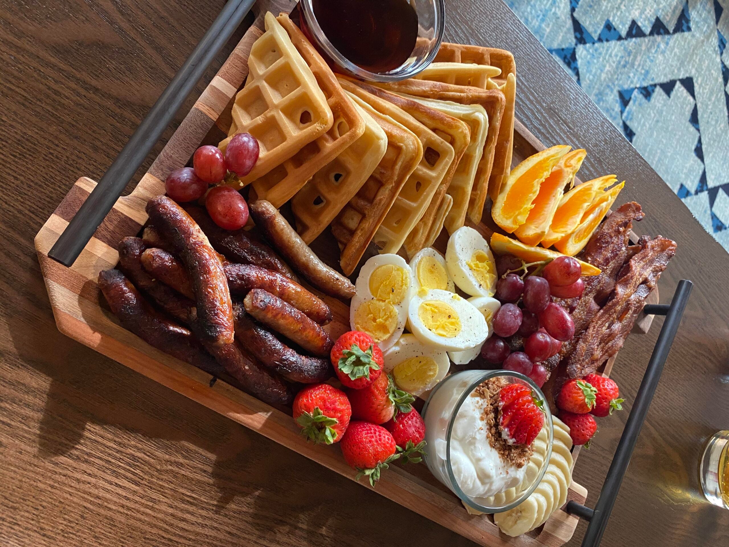 A breakfast platter on a wooden tray with waffles, sausages, hard-boiled eggs, bacon, sliced oranges, strawberries, grapes, banana slices, and a bowl of yogurt.