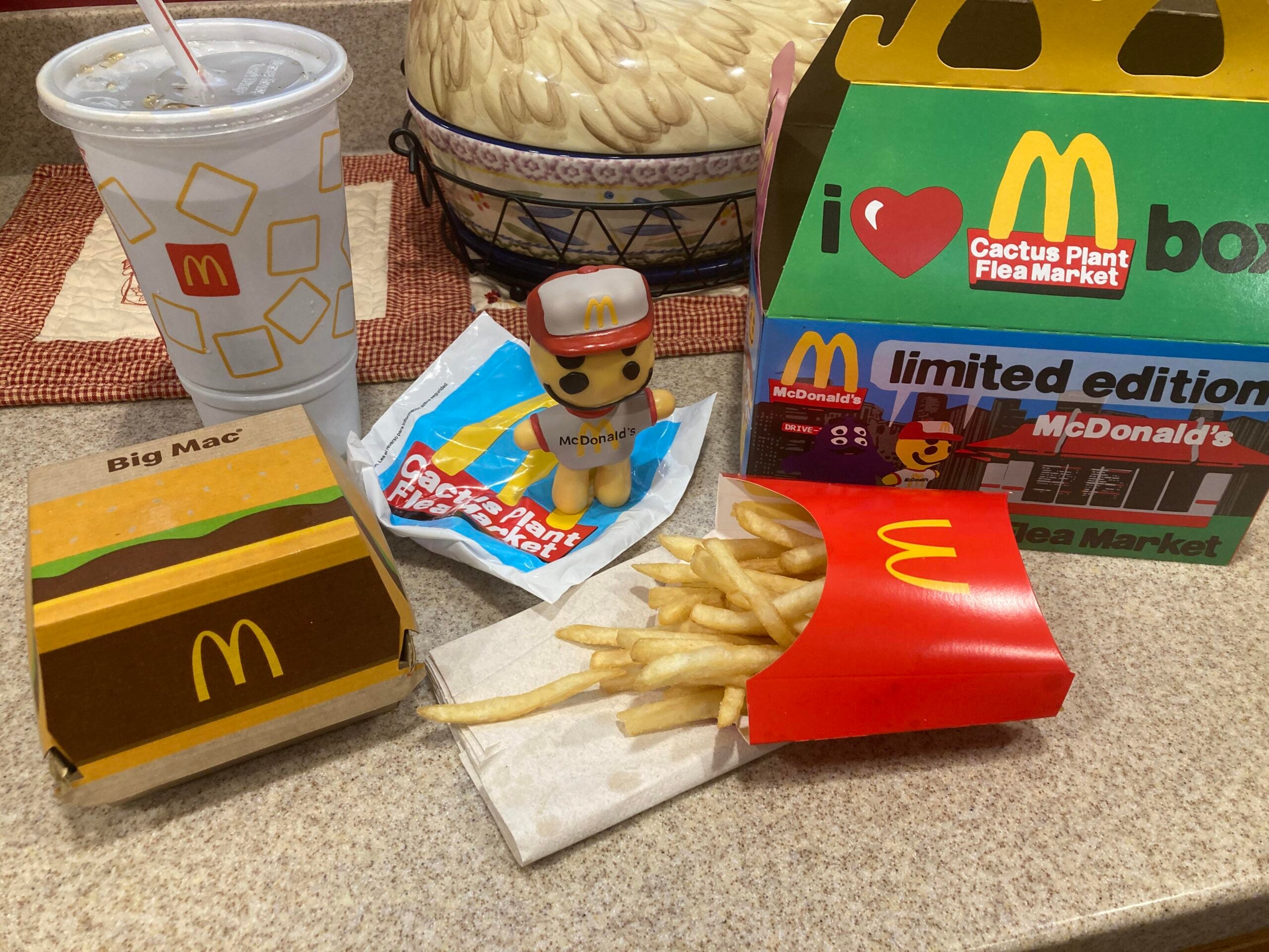A McDonald’s Cactus Plant Flea Market meal including a Big Mac box, a drink, a toy figurine, and a small container of french fries next to the promotional meal box, one of the fast-food prices.