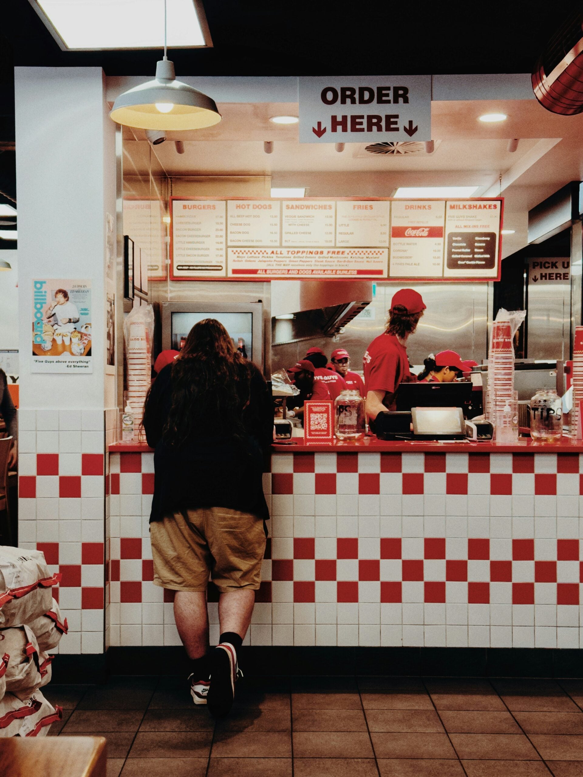 A person stands at the order counter of a fast-food restaurant with red and white tiles while employees work behind the counter. “Order Here” sign hangs above.