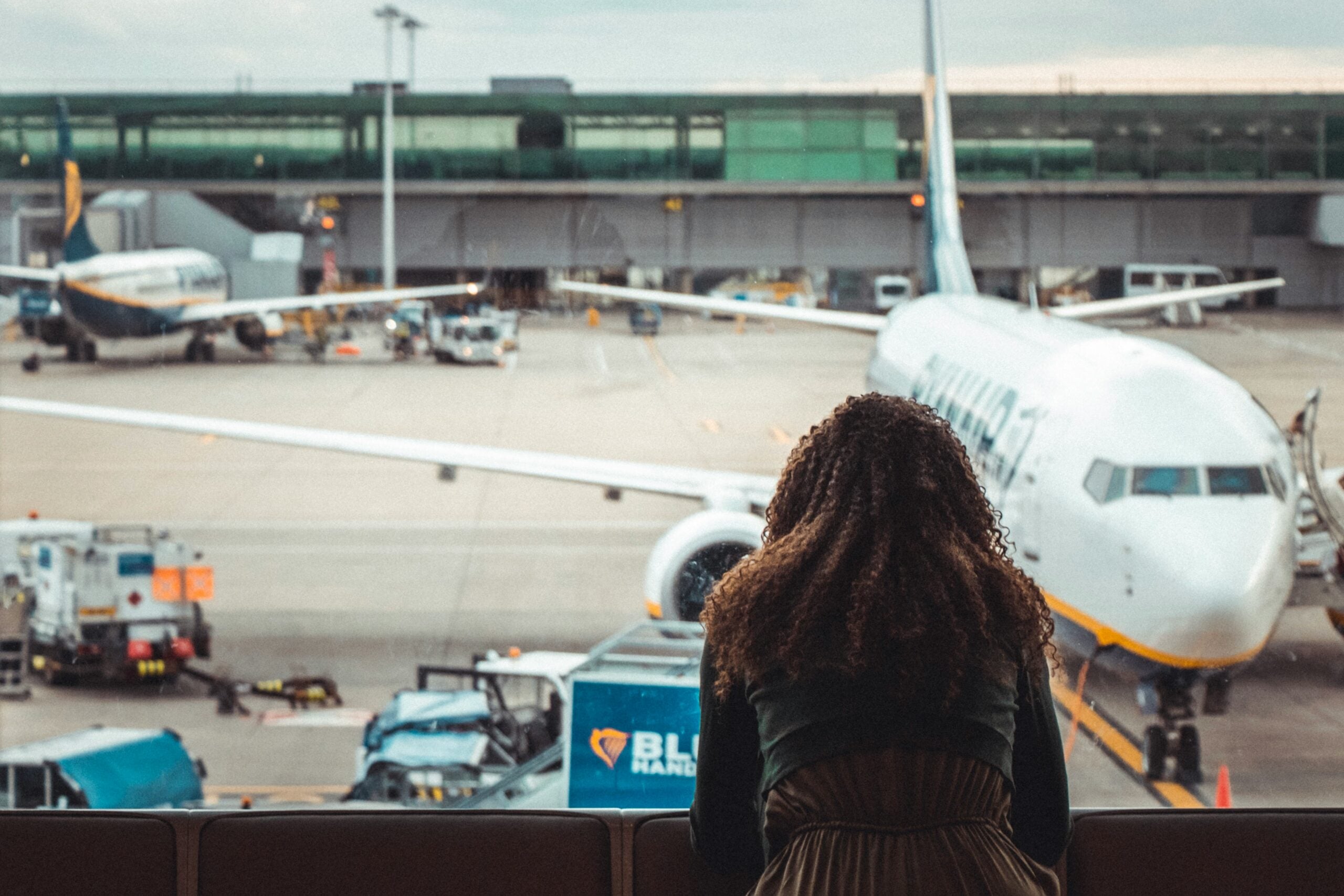 A person with long, curly hair looks out an airport window at airplanes parked on the tarmac.