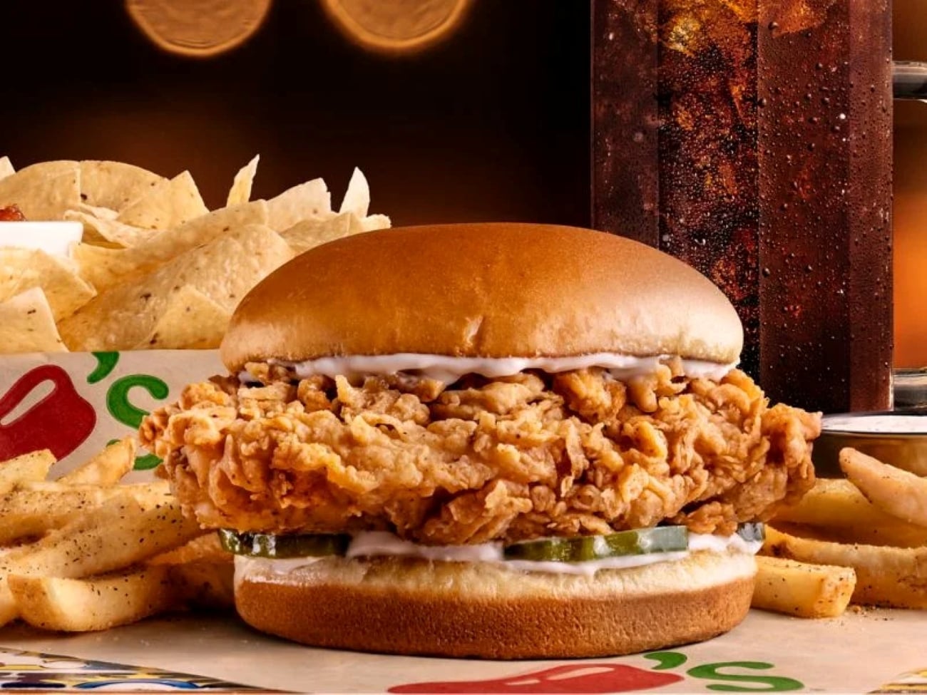 A fried chicken sandwich with pickles on a bun, served with a side of fries, tortilla chips, and a drink.