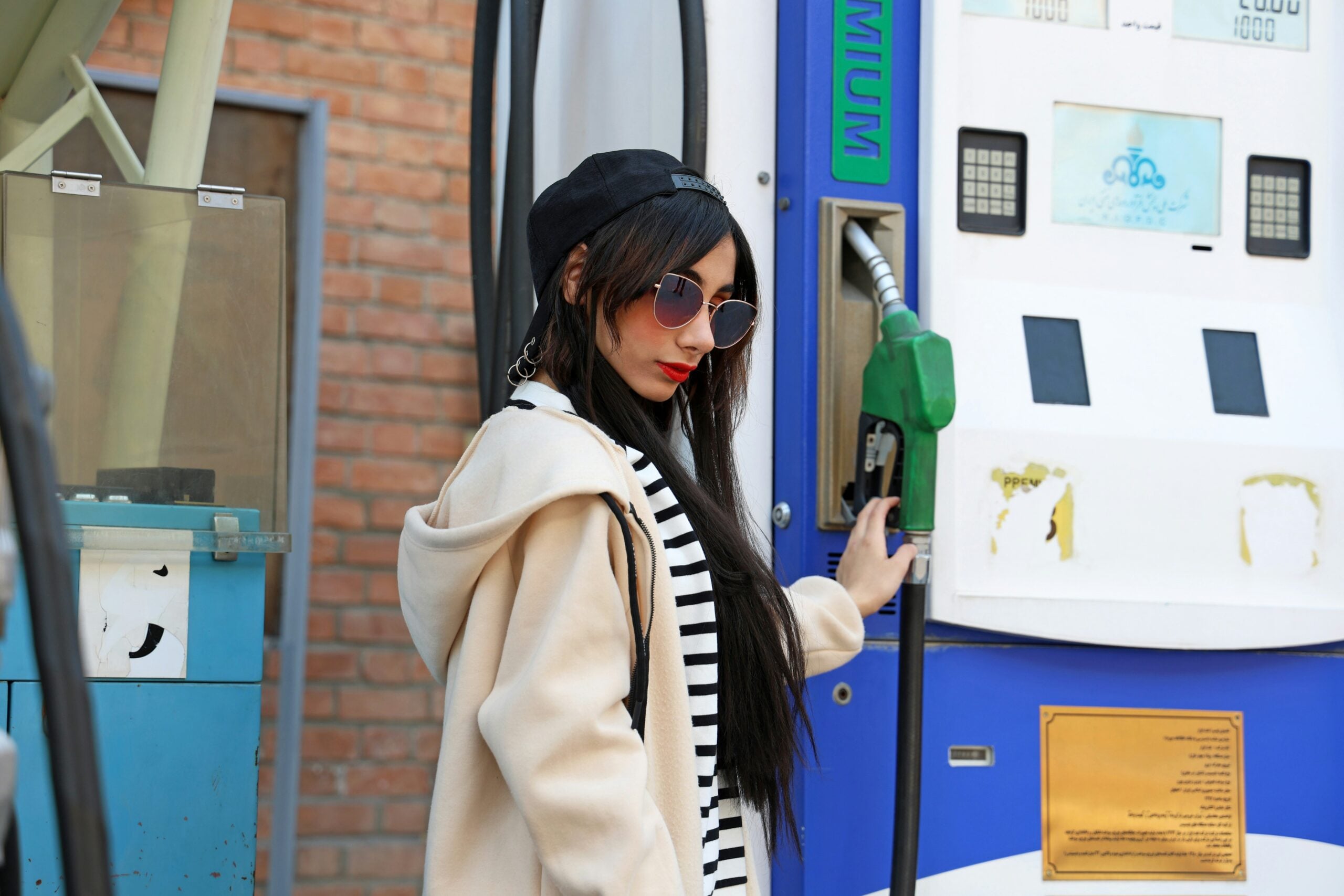 A woman in a cream-colored jacket, striped shirt, and black baseball cap reaching for a green gas pump nozzle at a station.