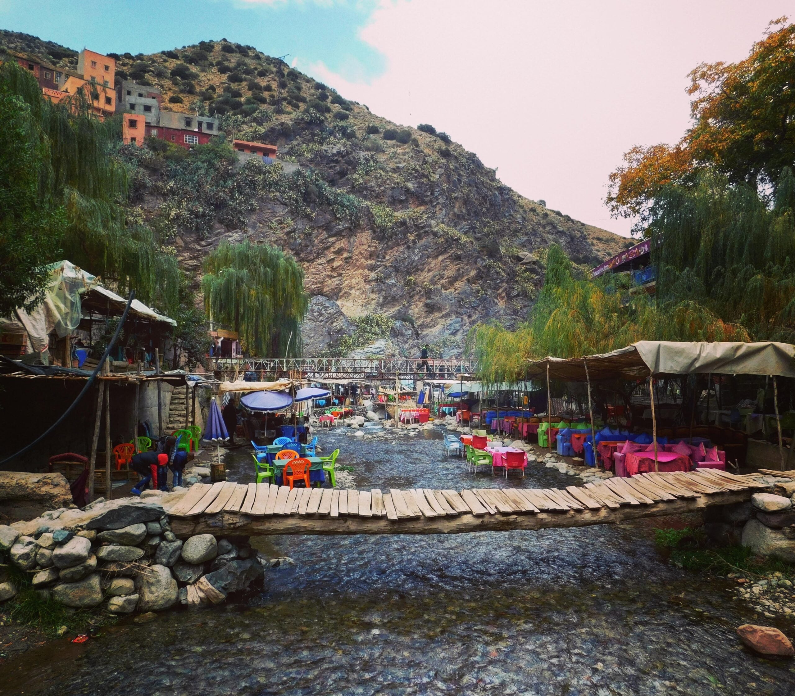 A rustic wooden footbridge crosses a shallow stream, with colorful outdoor tables and chairs on both sides, surrounded by trees and hills with houses in the background.