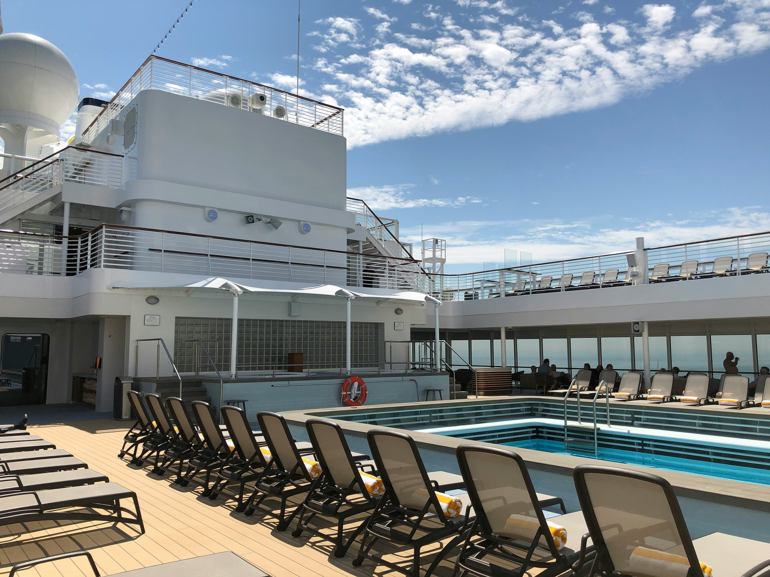 Rows of lounge chairs line a cruise ship deck next to a rectangular swimming pool under a sunny sky.