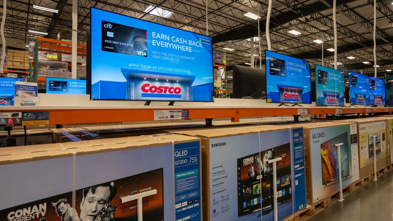 A row of flat-screen televisions on display shelves in a retail store, each showing a Costco Visa credit card advertisement on screen.