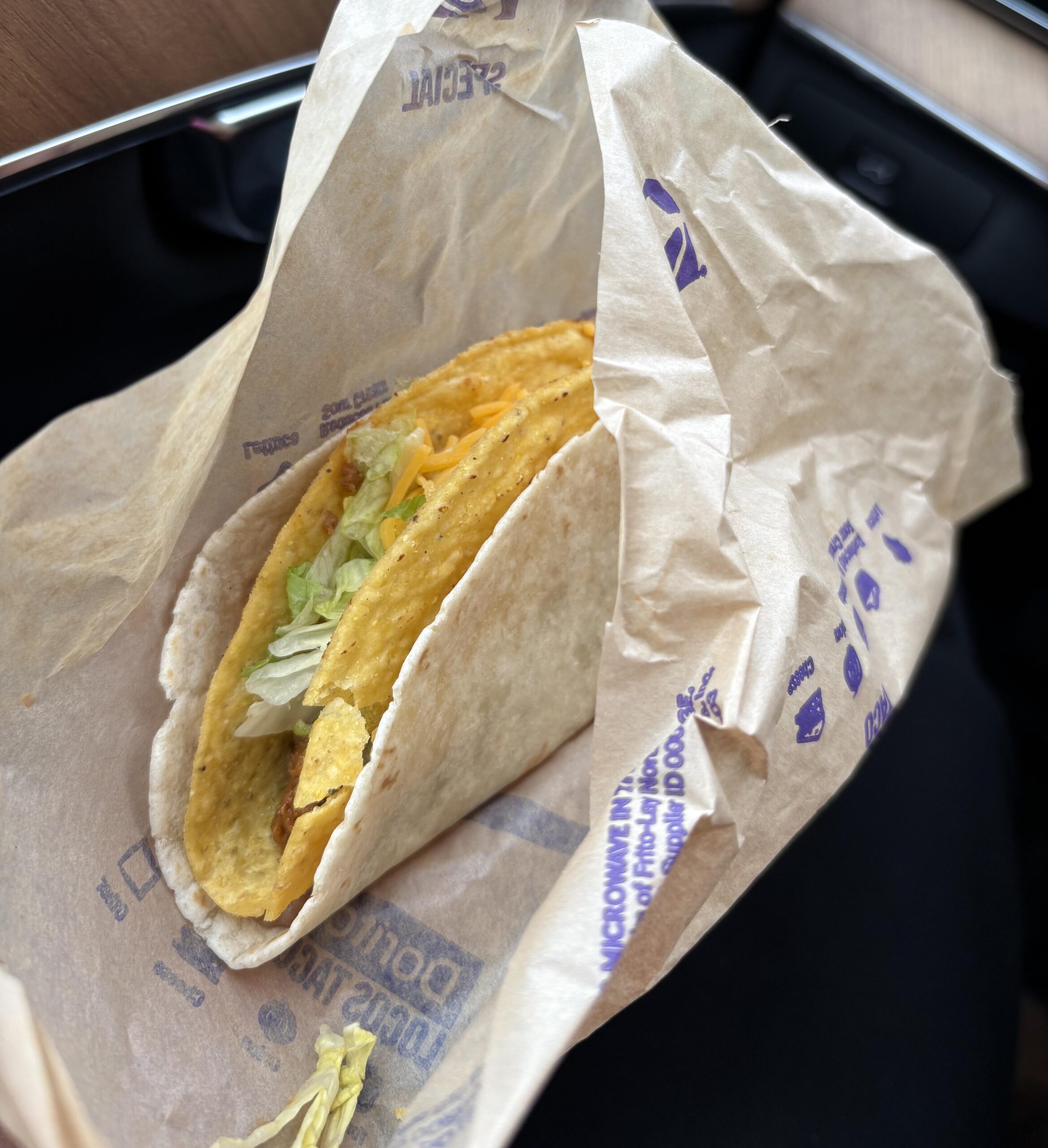 A crunchy taco with meat, lettuce, and shredded cheese inside a soft flour tortilla, held in a branded paper wrapper.