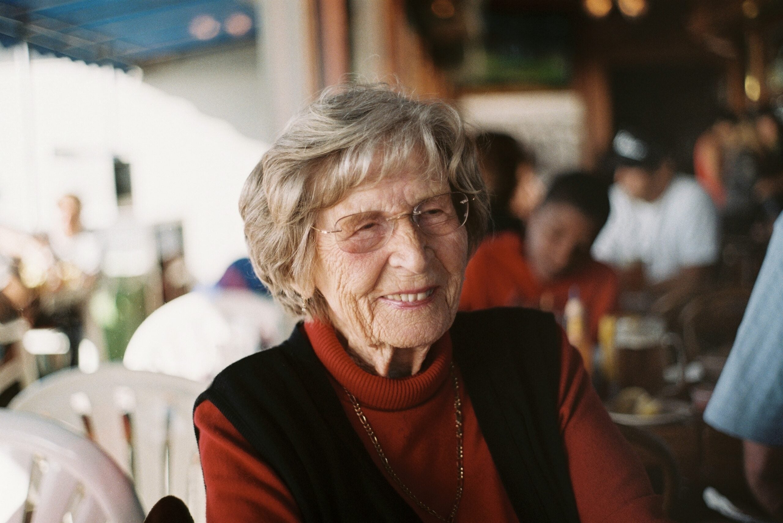 Elderly woman with glasses and short gray hair smiles while sitting indoors at a busy restaurant, wearing an orange sweater and black vest.
