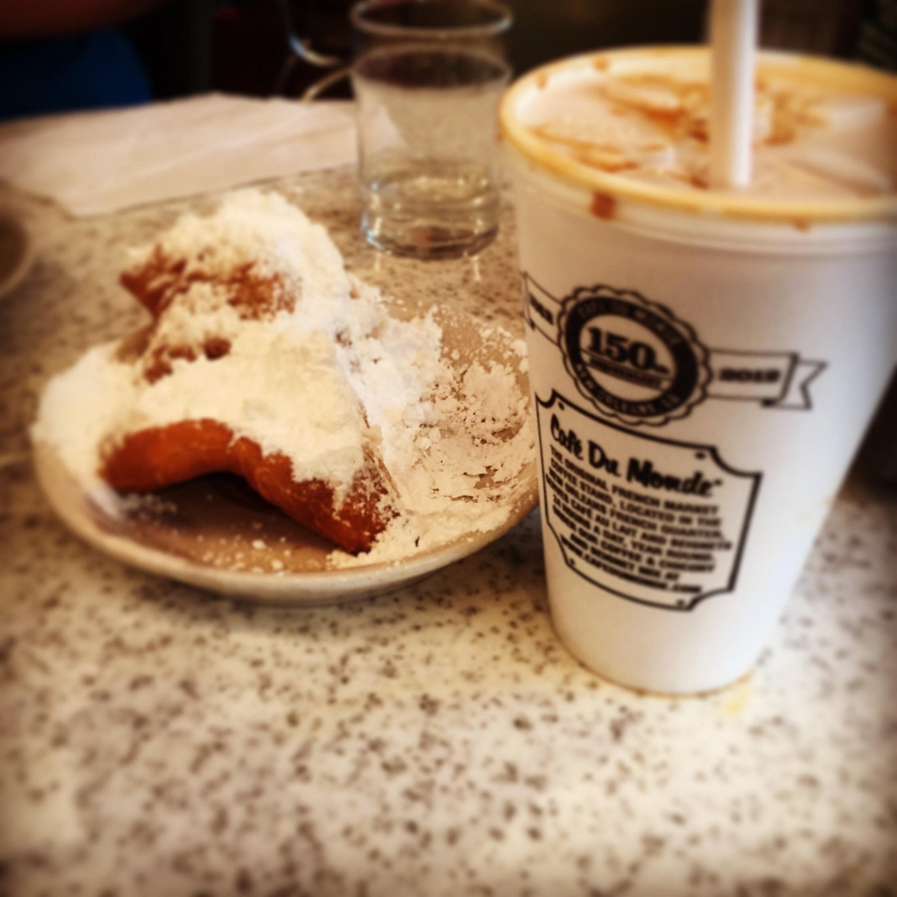 A beignet covered in powdered sugar on a small plate next to a Café Du Monde iced coffee in a branded paper cup.