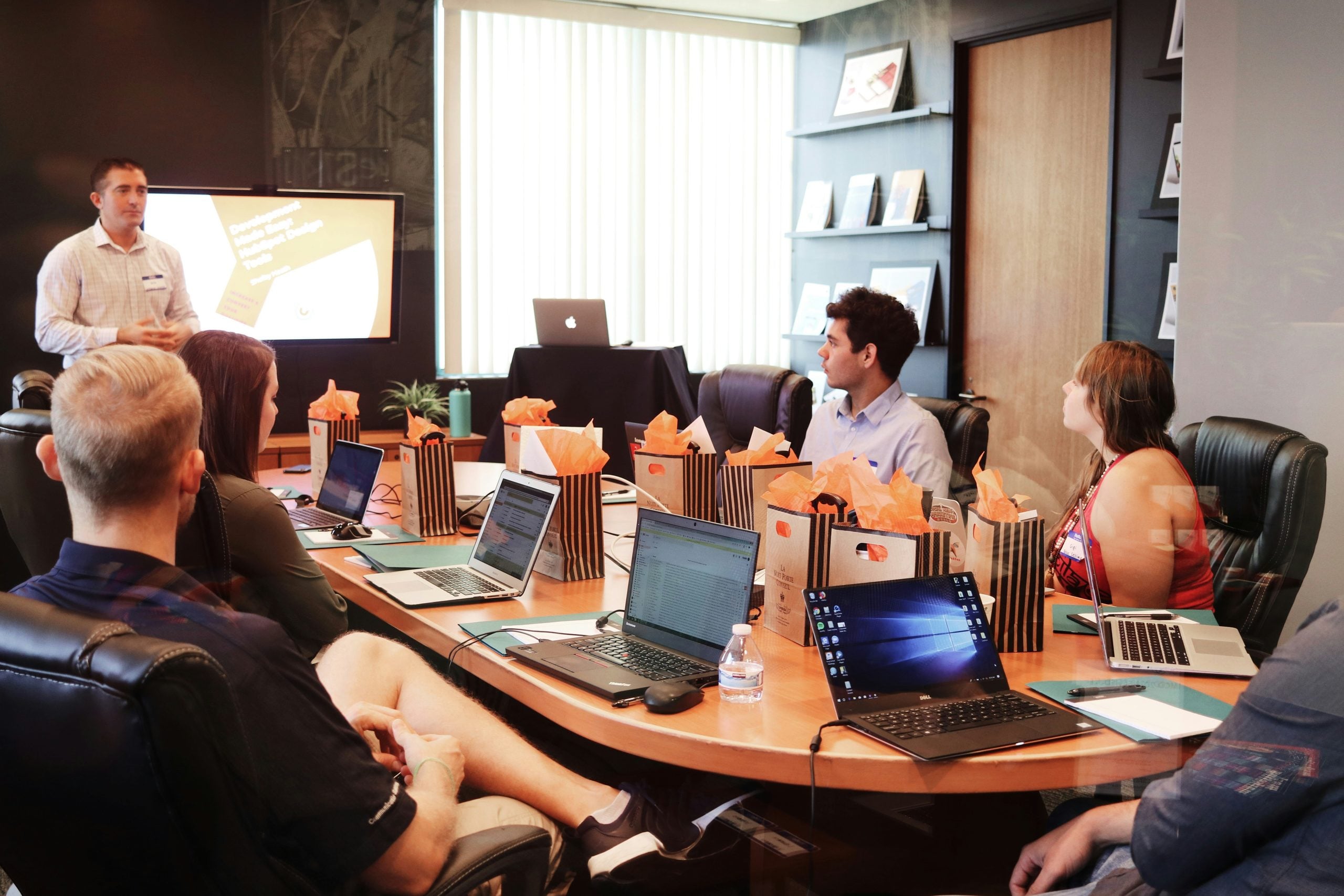 A group of people sit around a conference table with laptops, listening to a presenter standing near a screen. Gift bags are on the table. The room has large windows and bookshelves.