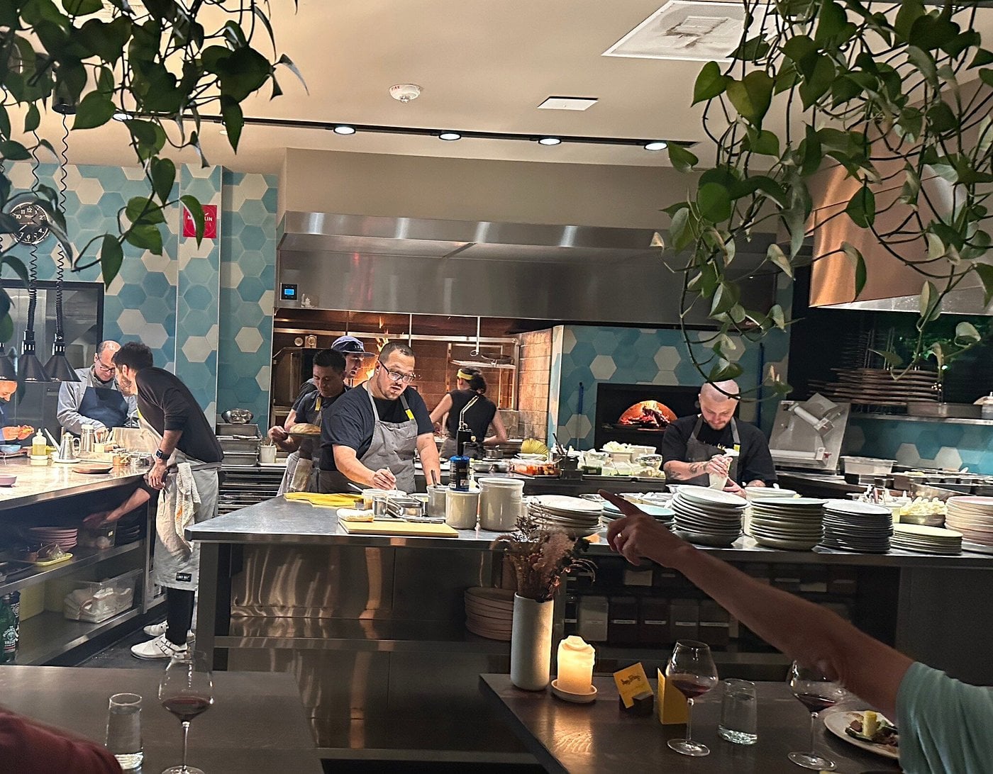 Chefs prepare food at the bar seating in an open kitchen behind a stainless steel counter, with a wood-fired oven visible in the background.
