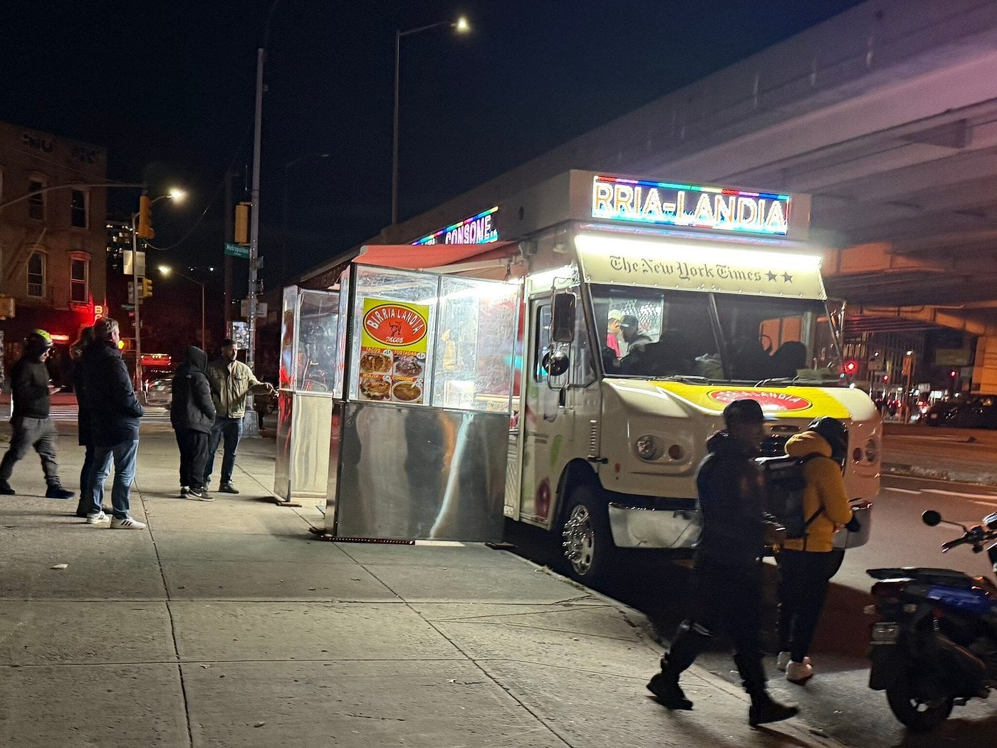 A Birria-Landia food truck parked on a city sidewalk at night with customers waiting in line.