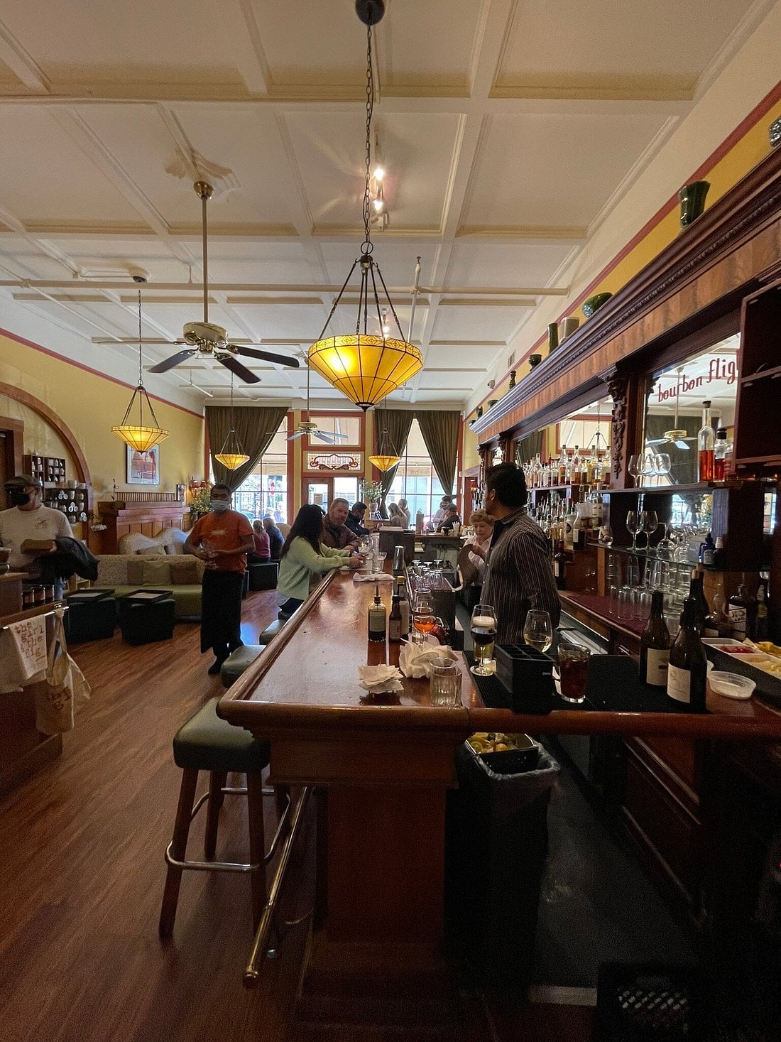 People sitting at a long wooden bar in a restaurant with high ceilings and pendant lights.