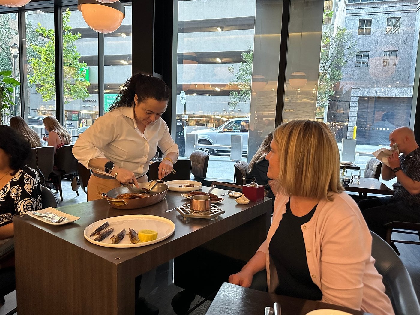 A server uses tongs to portion food from a copper pan onto a plate for a diner at a restaurant table.