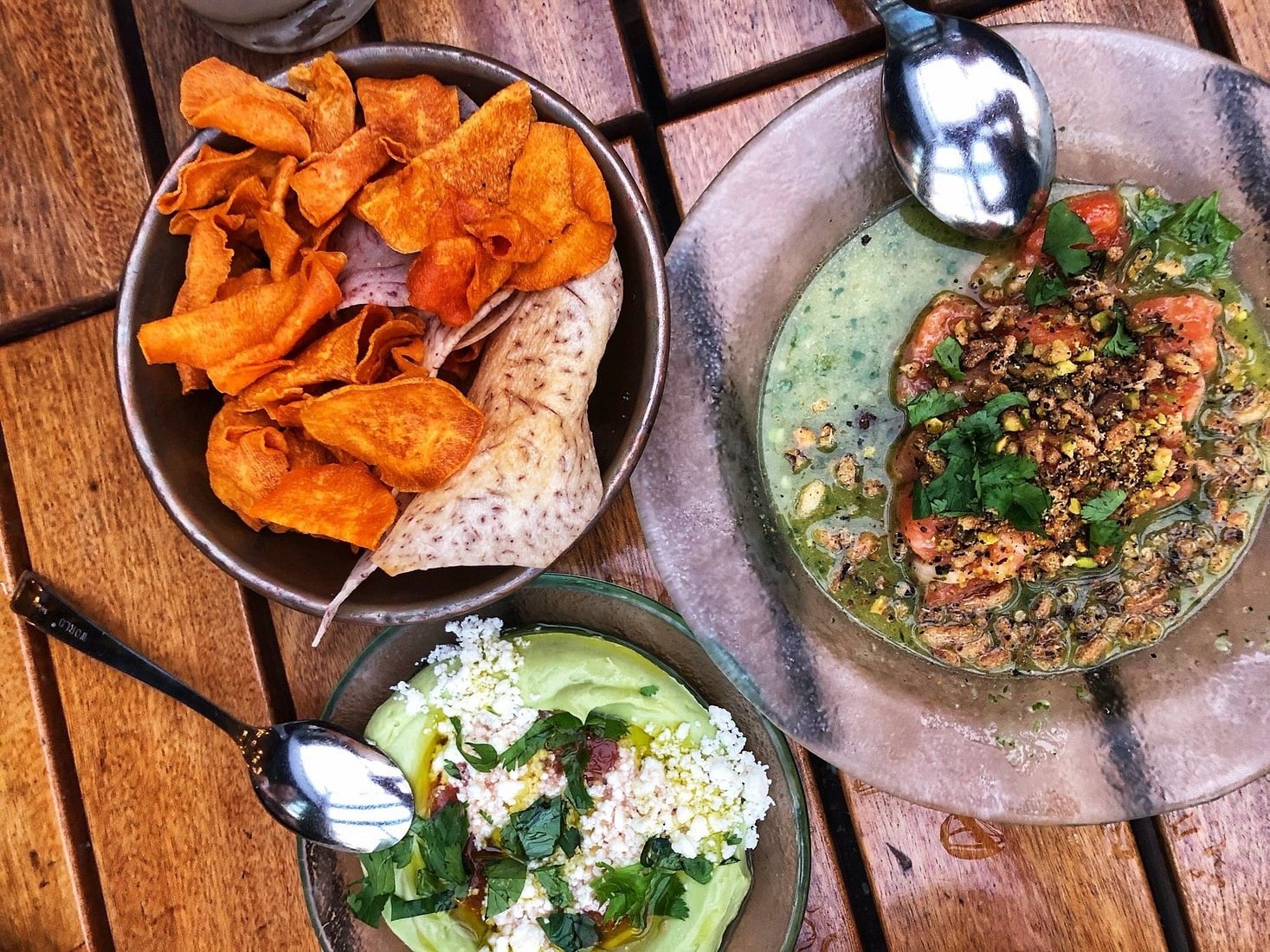 A bowl of root vegetable chips, a bowl of guacamole topped with crumbled cheese and herbs, and a bowl of ceviche topped with herbs and nuts, arranged on a wooden table.