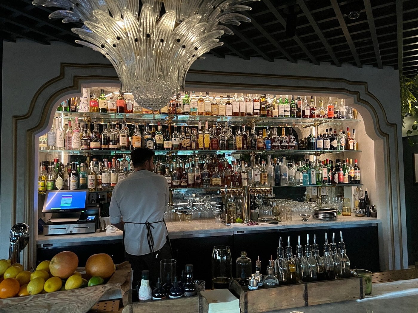 A bartender stands behind a bar with a multi-tiered back display of liquor bottles and a large, decorative glass chandelier hanging overhead.