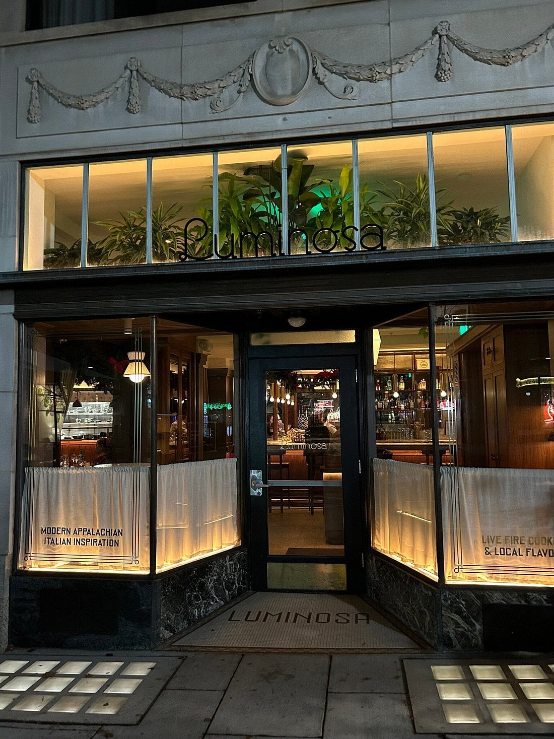 The storefront of a restaurant named Luminosa, featuring glass windows with the text “Modern Appalachian Italian Inspiration” and “Live Fire Cooking & Local Flavors.”