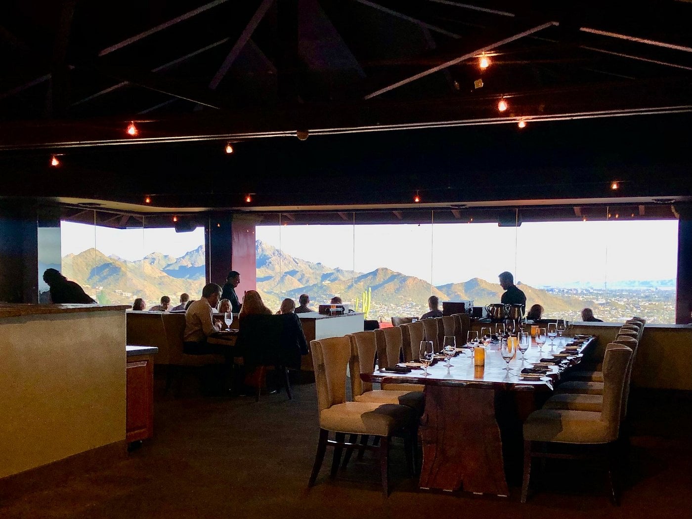 Diners seated at tables in a restaurant overlooking a desert mountain landscape at sunset.