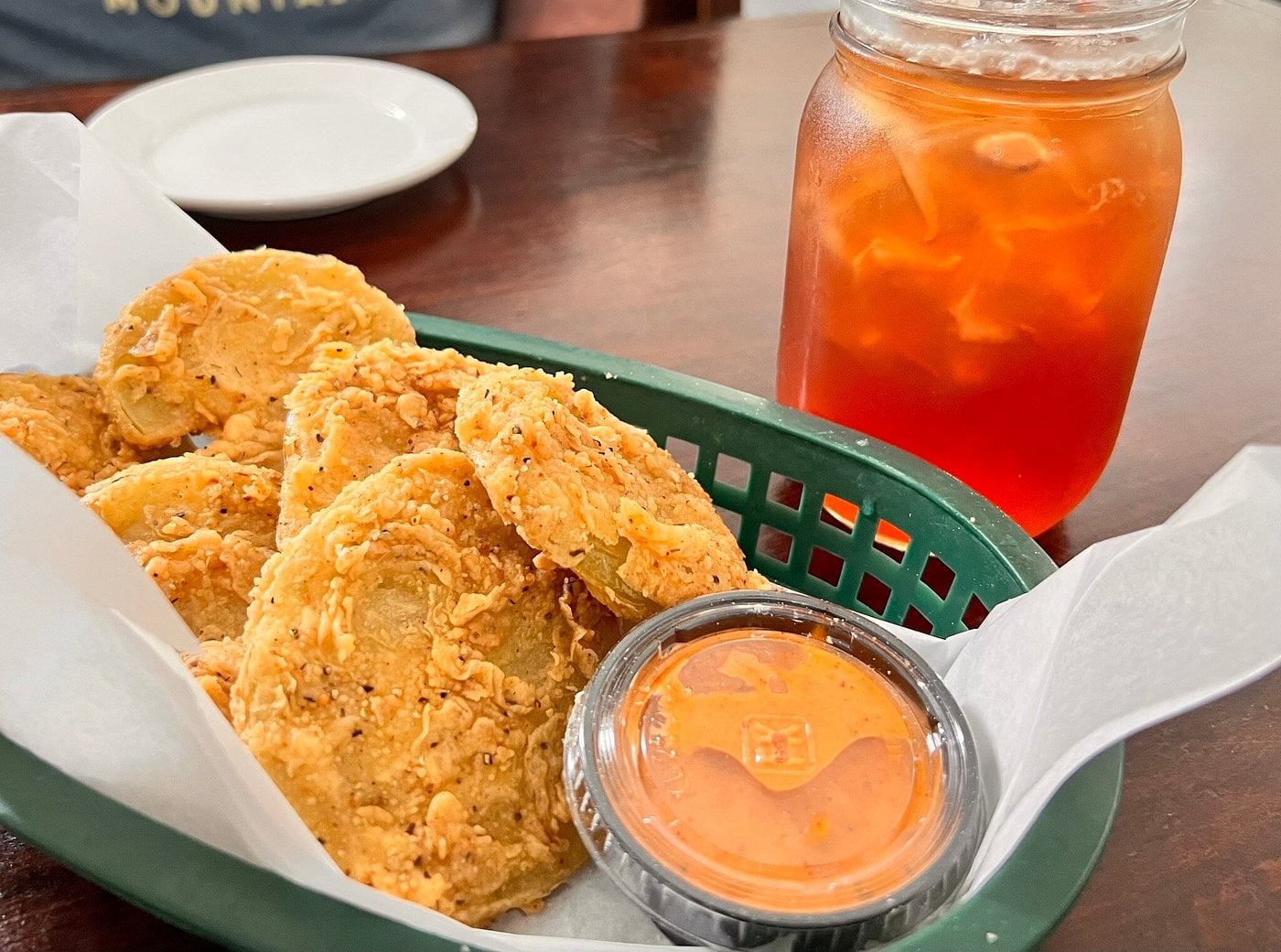 A basket of fried green tomato slices with a side of dipping sauce and a glass of iced tea.
