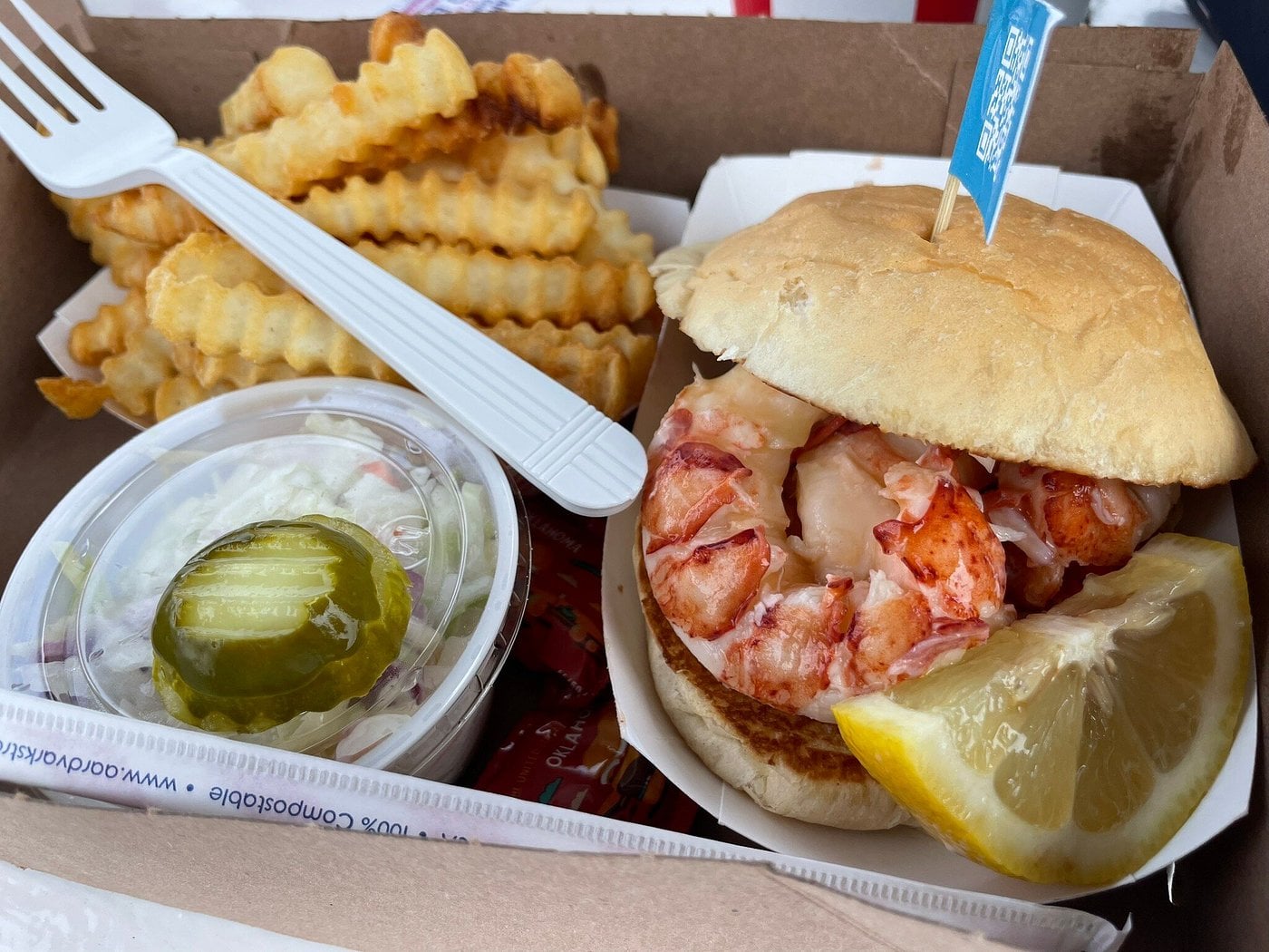 A cardboard takeout box containing a lobster roll, a side of crinkle-cut fries, and a container of coleslaw topped with a pickle slice.