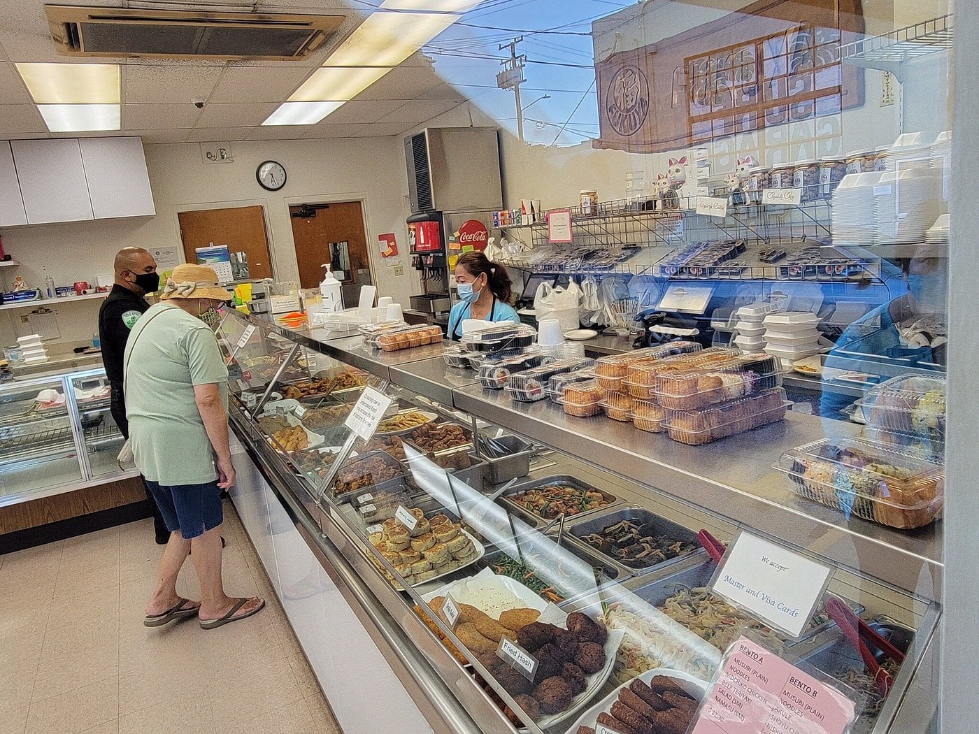 A customer in a straw hat stands at a deli counter viewing prepared foods while an employee in a blue shirt and face mask works behind the glass.
