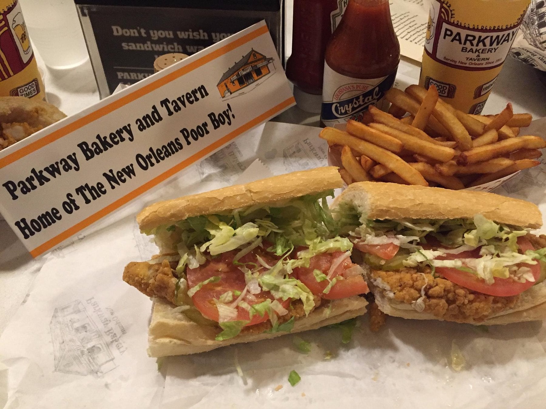 A fried seafood po’ boy sandwich cut in half on parchment paper, served with a side of french fries next to a Parkway Bakery and Tavern promotional card and a bottle of Crystal hot sauce.