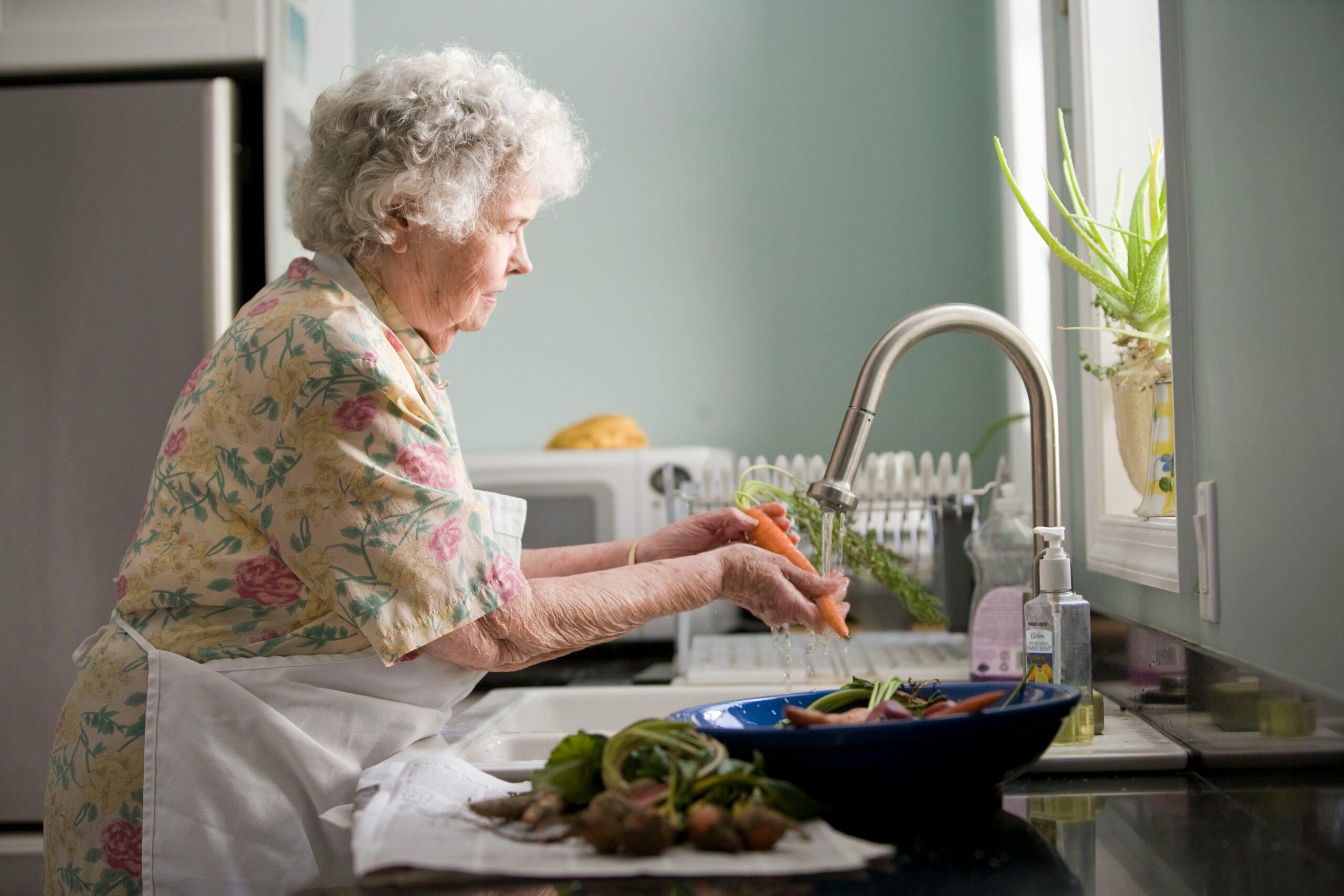 An elderly woman wearing a floral dress and white apron washes carrots at a kitchen sink, with more vegetables and a blue bowl on the counter beside her. Sunlight comes in through a window above the sink.