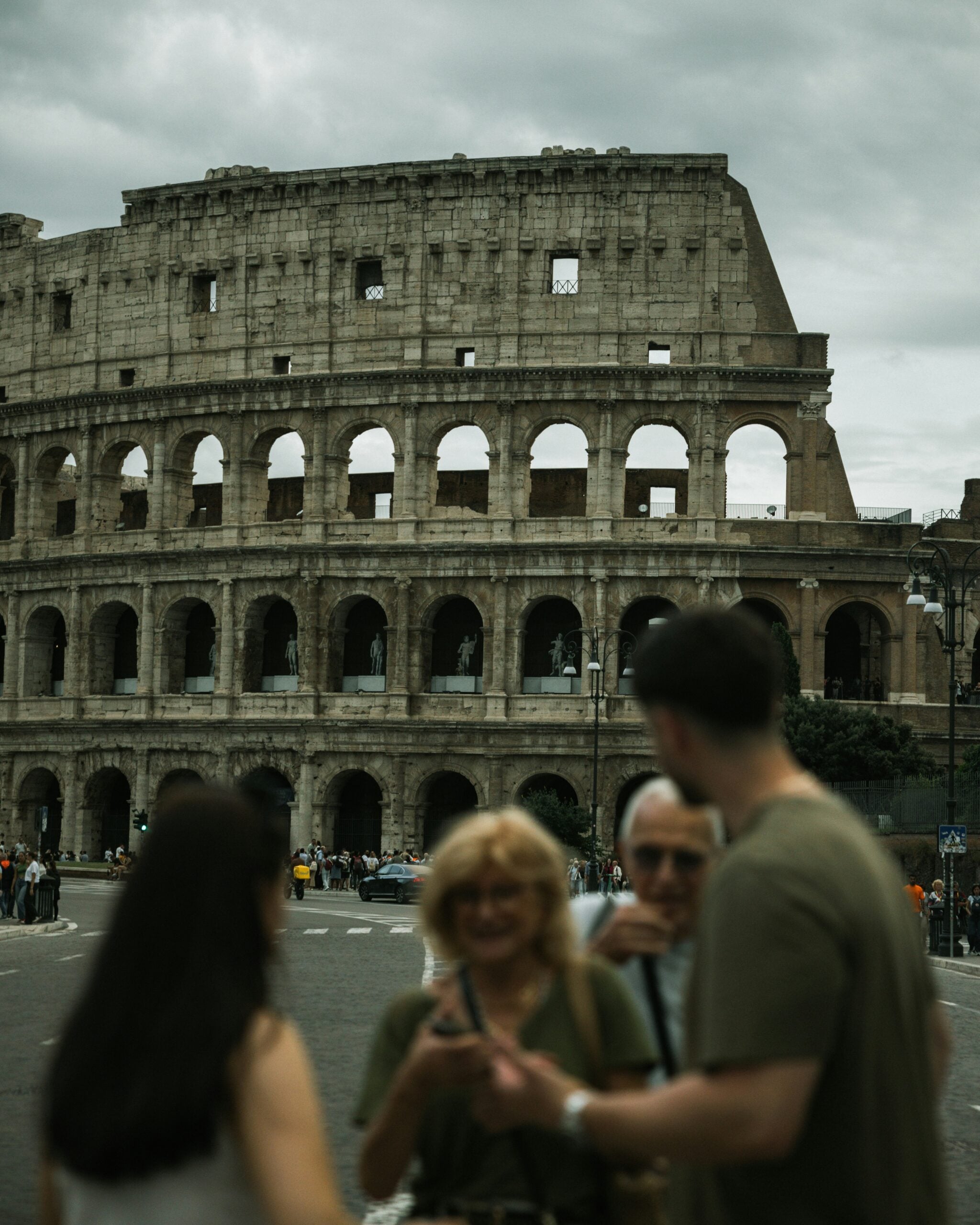 Four people are standing in the foreground, out of focus, with the ancient Roman Colosseum in sharp focus behind them under a cloudy sky. The area is busy with other tourists in the background.