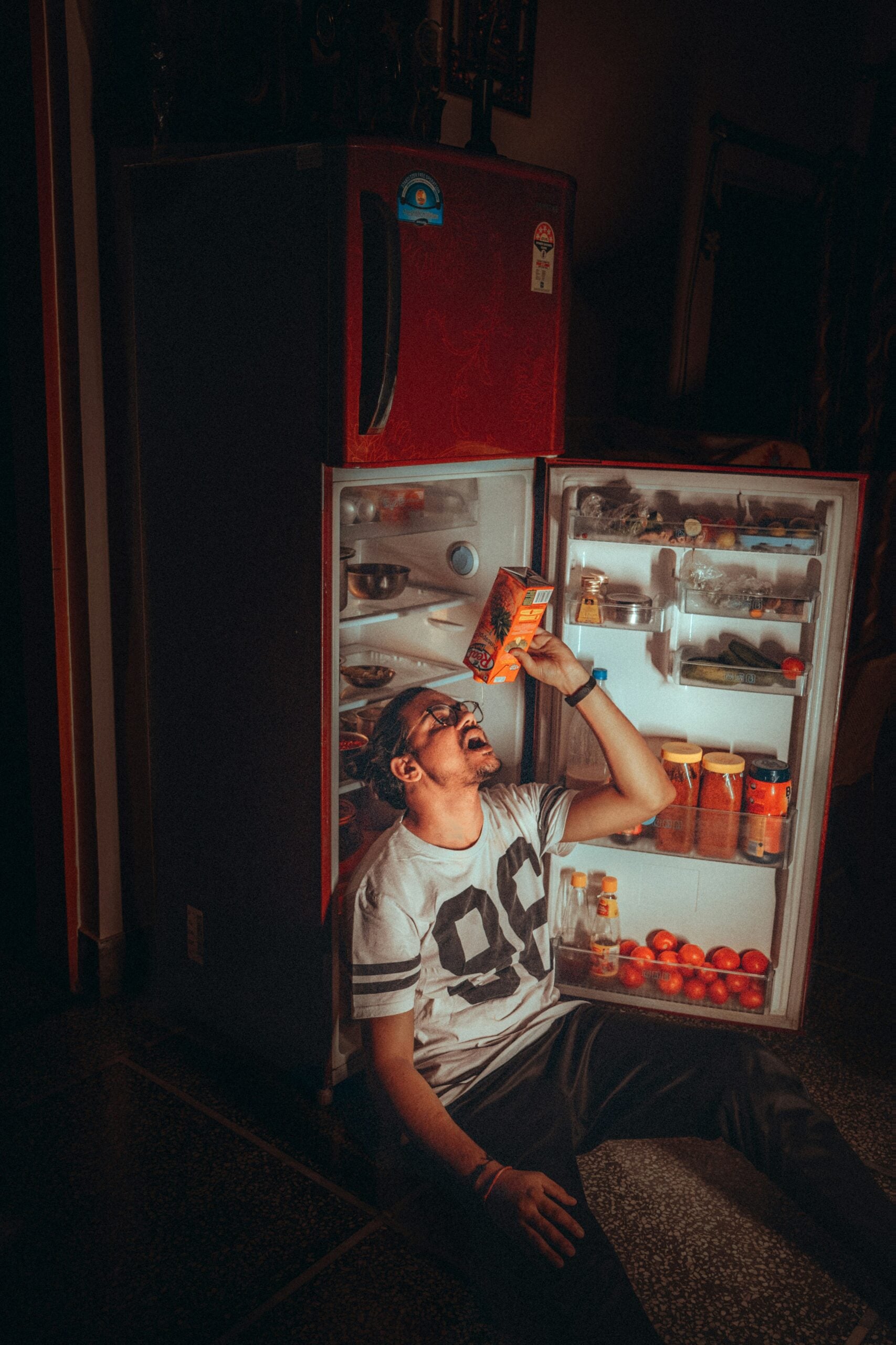 A man sitting on the floor in front of an open refrigerator, tilting his head back to drink from a carton of juice.