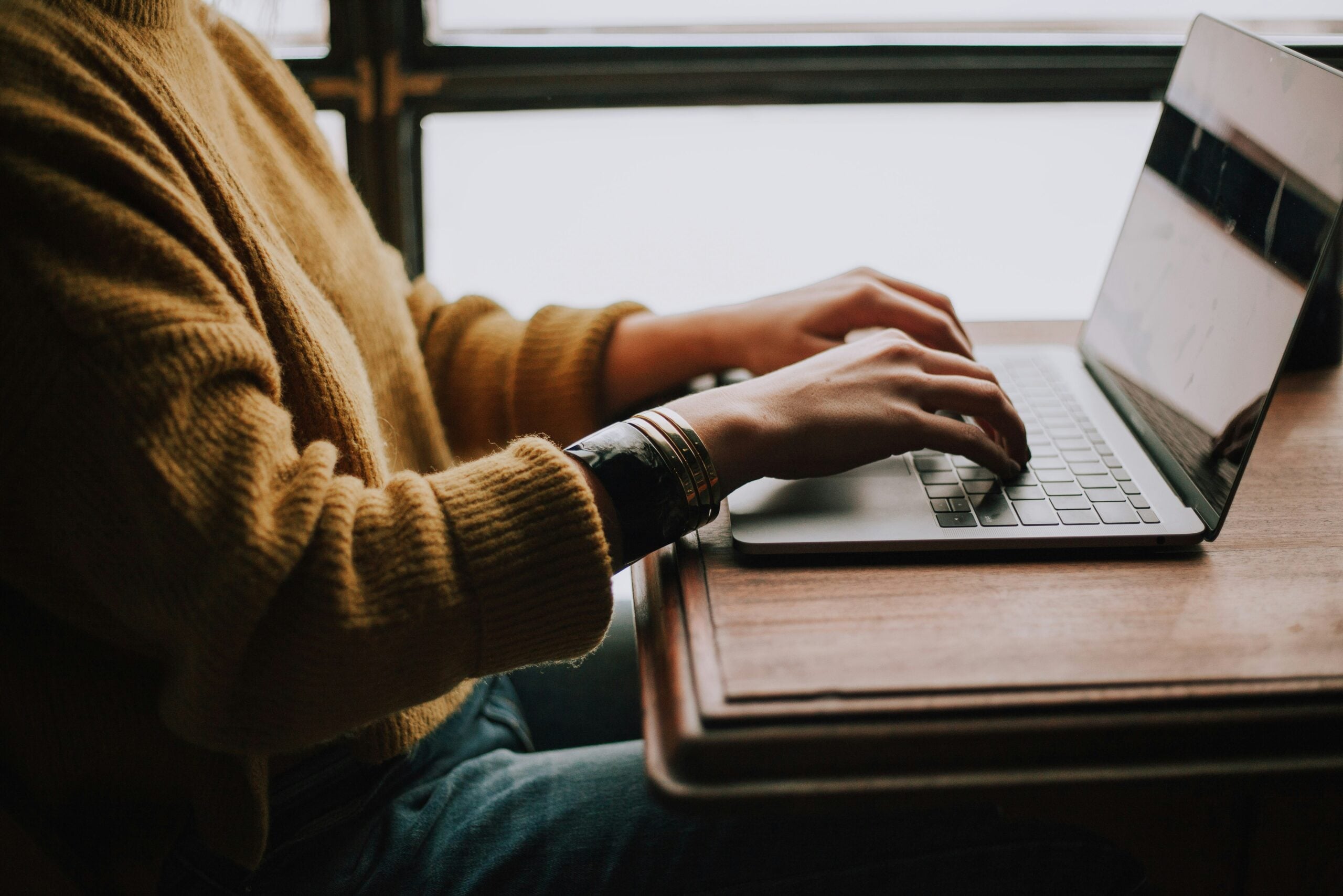 A person in a yellow sweater types on a laptop at a wooden table.