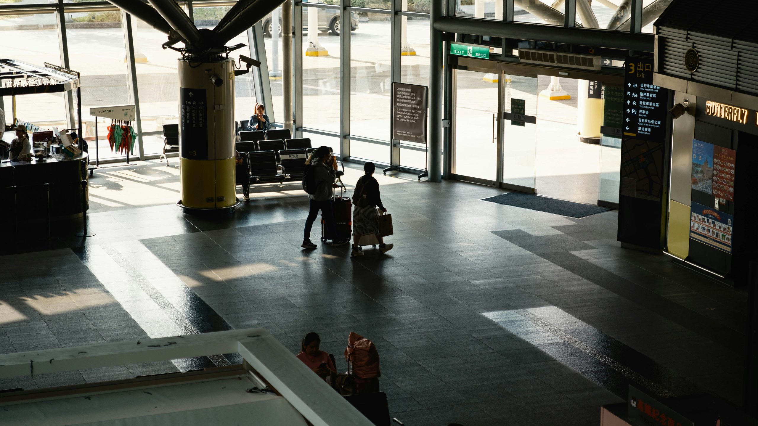 Two people with suitcases walk through a spacious, sunlit airport terminal. Shadows are cast on the floor, and there are seats, signs, and large windows showing the outside area.