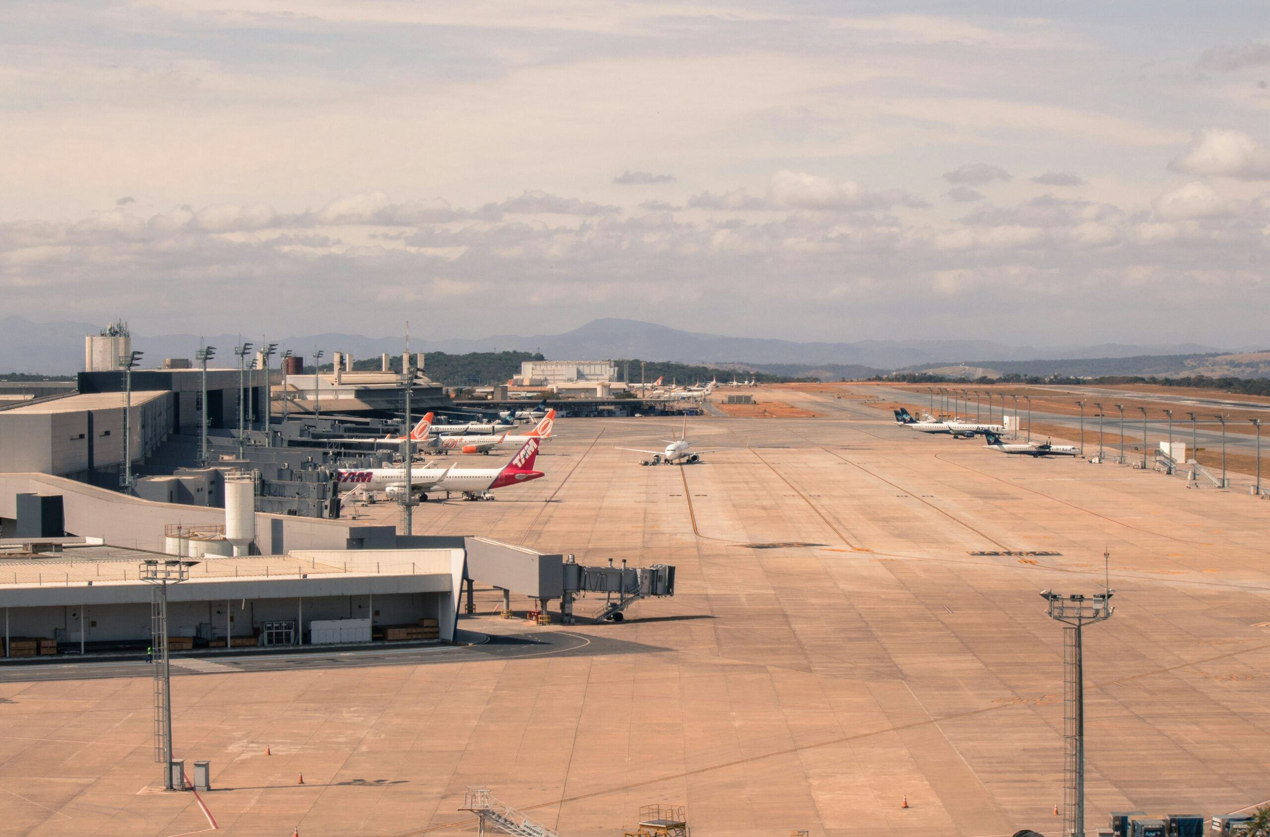 Several passenger airplanes parked at airport gates with a runway and distant hills in the background.