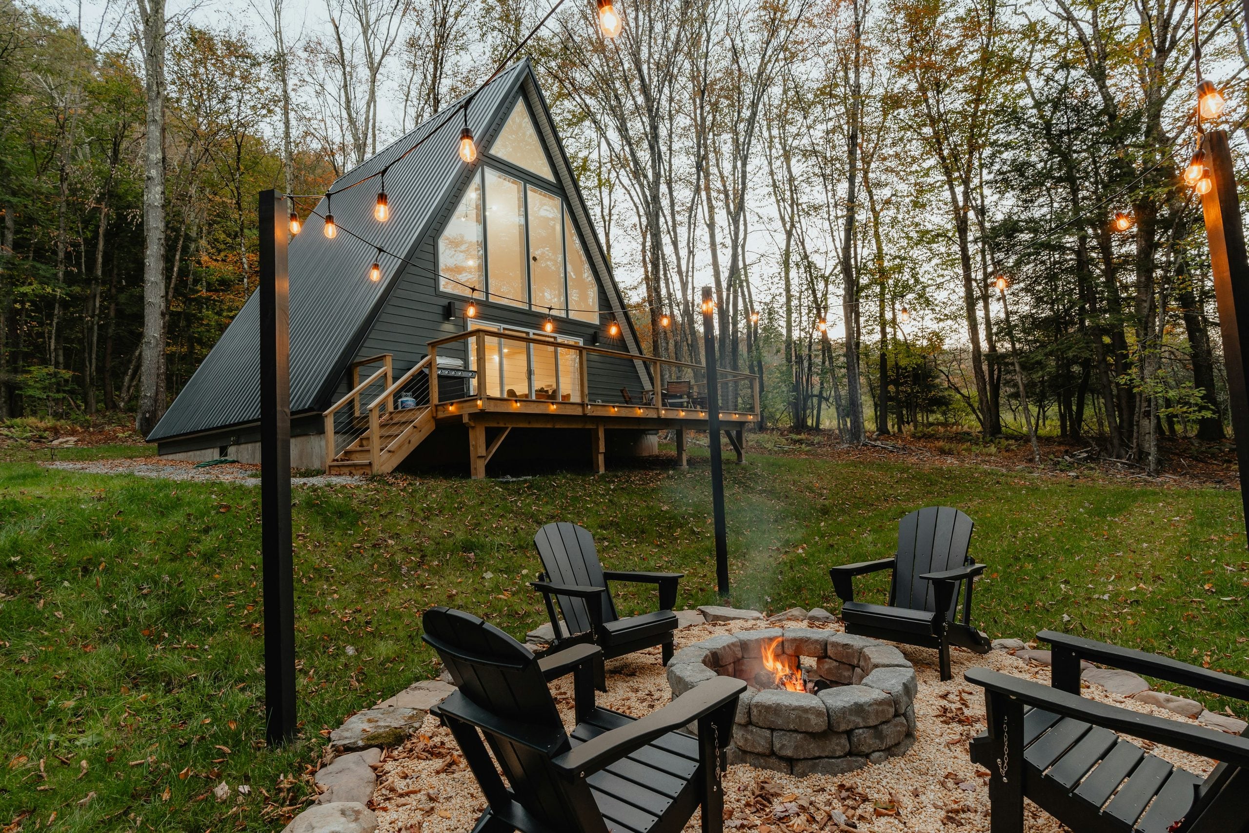 A modern A-frame cabin with large windows sits in a wooded area. Four black Adirondack chairs surround a stone fire pit with string lights above, creating a cozy outdoor setting.