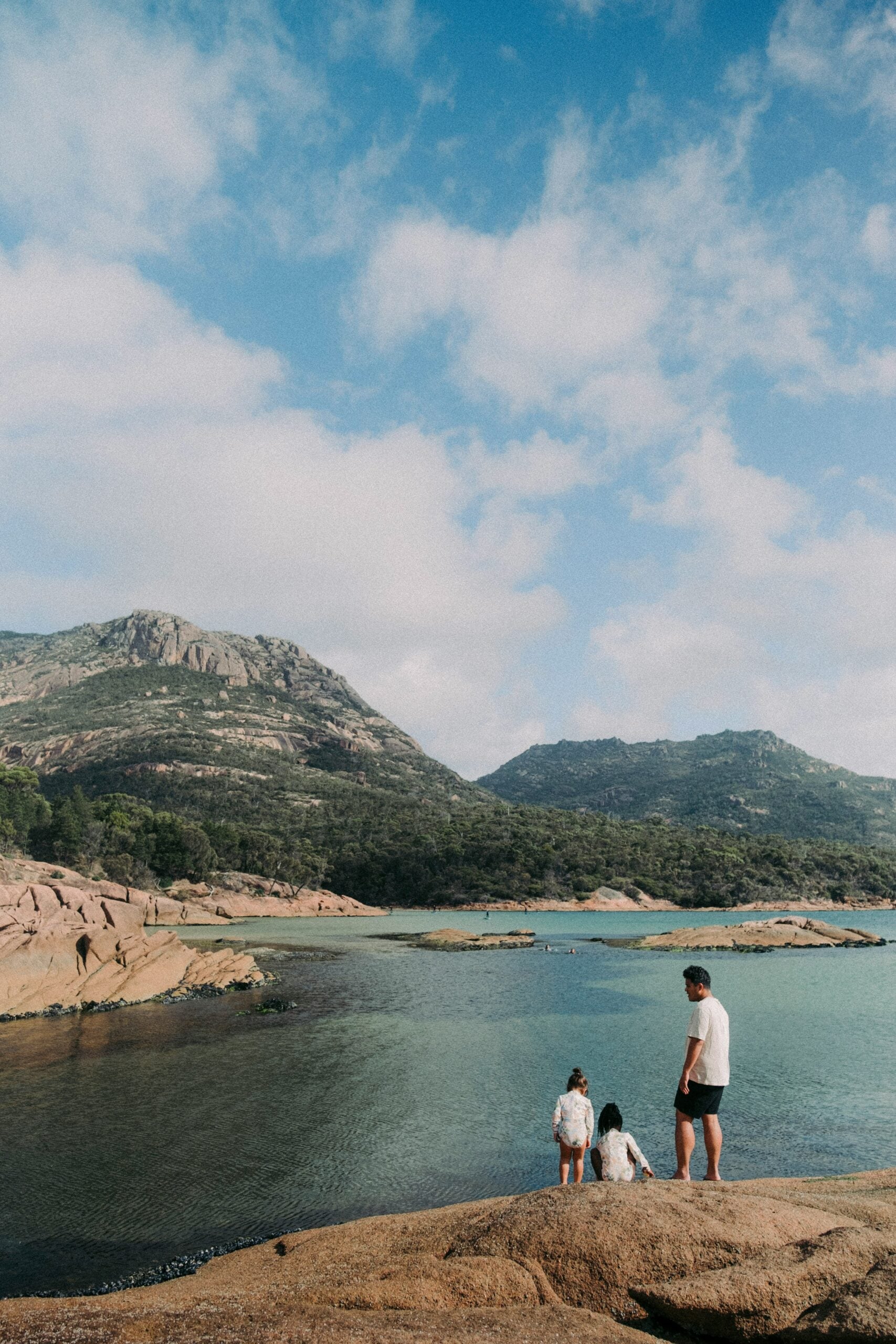 Three people stand on rocky terrain beside clear blue water, with green mountains and a partly cloudy sky in the background.