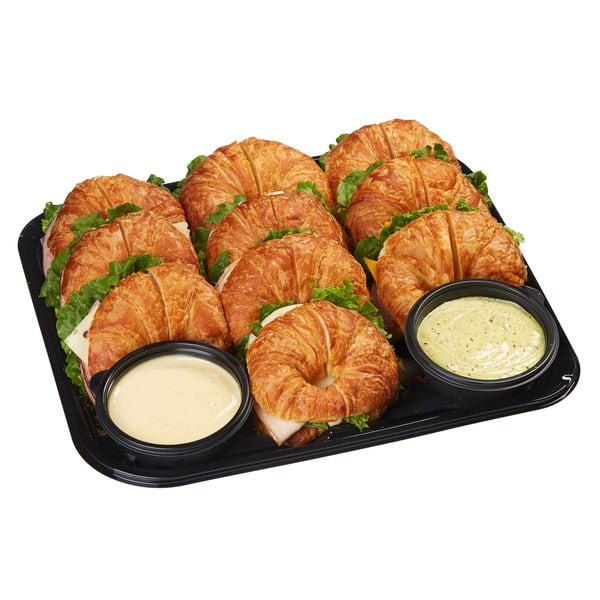 A black catering tray filled with several croissant sandwiches, lettuce, and two small bowls of dipping sauces.